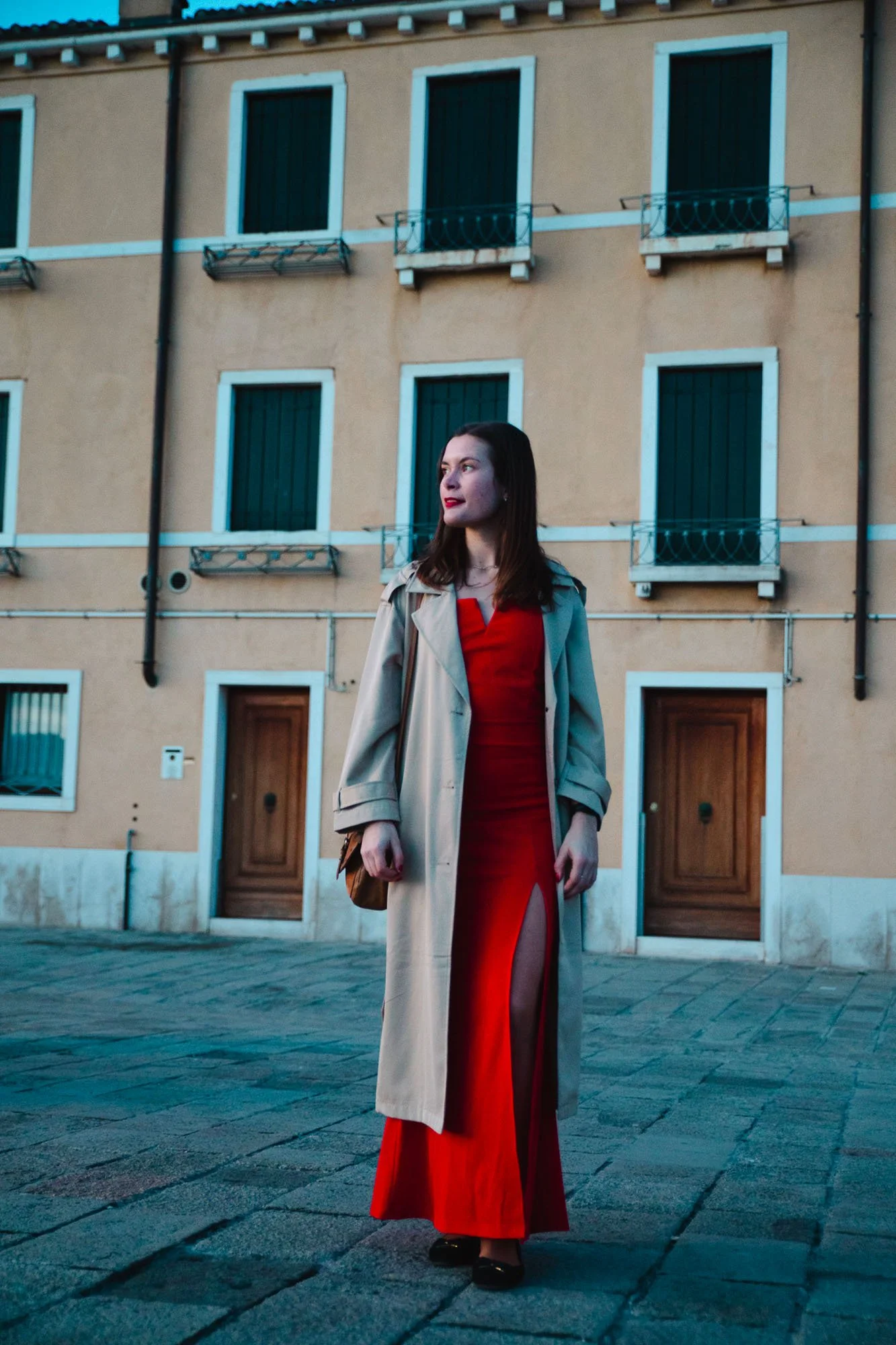 Donna con abito rosso lungo con slit davanti, cappotto beige e borsa marrone, in piedi davanti a un palazzo con finestre con inferriate blu in una scena urbana serale.