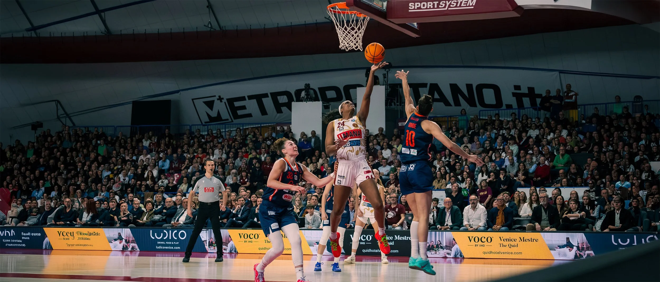 Partita di basket femminile in corso con giocatrici che si contendono il pallone sotto canestro, pubblico numeroso sugli spalti, arbitro osserva, ambiente sportivo interno.