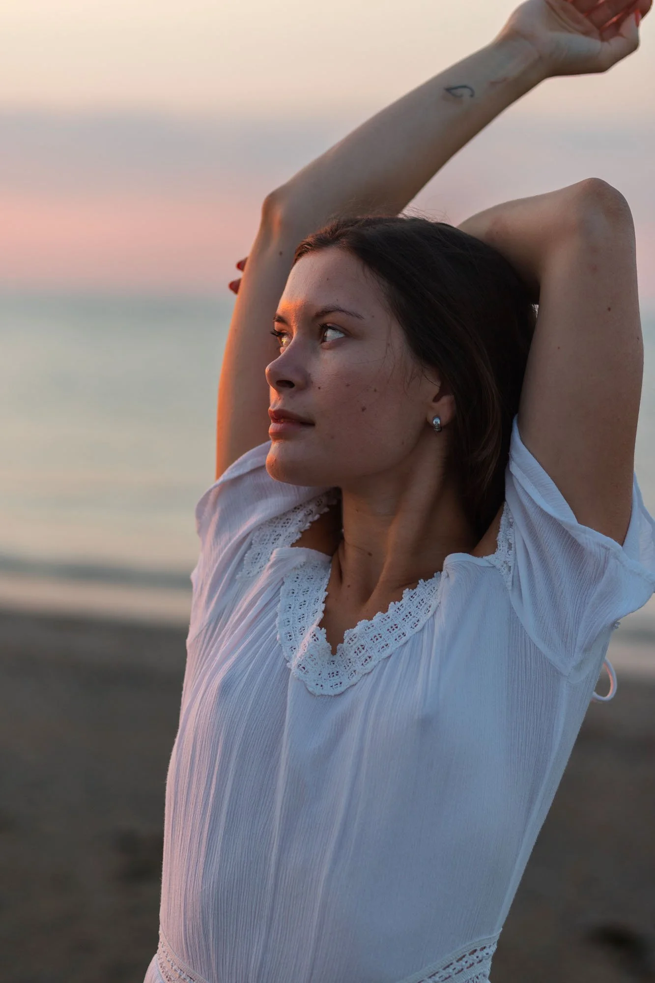 Una donna con la pelle chiara e i capelli castani chiusi, indossa una camicia bianca con dettagli in pizzo. Sfondo sul mare al tramonto. È in posa con le braccia sollevate sopra la testa, guardando verso sinistra.