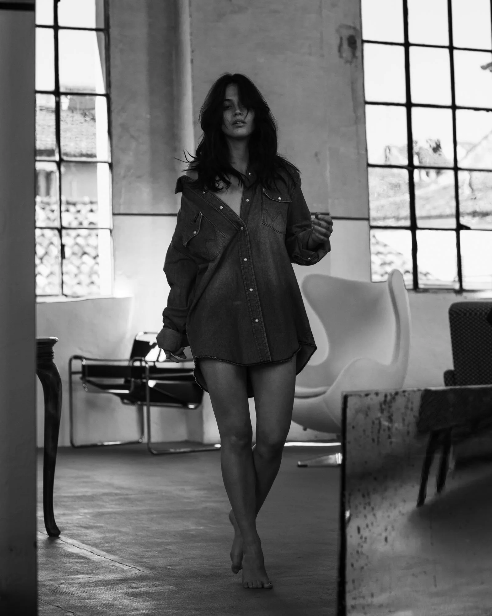 Ragazza con lunghi capelli scuri, vestita con una camicia e un vestito, cammina a piedi nudi in una stanza con grandi finestre. La scena è in bianco e nero.