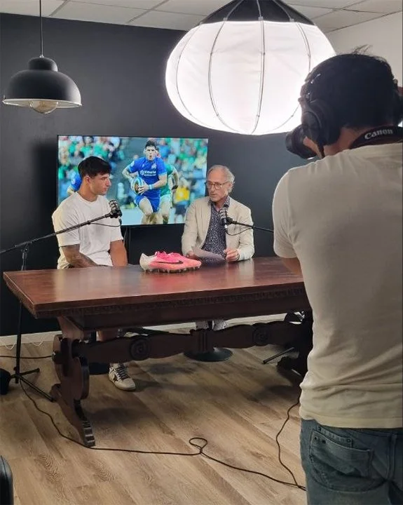 Stanza di registrazione di un podcast con tre uomini: uno che sta davanti al microfono, uno che sta presentando e un cameraman con videocamera