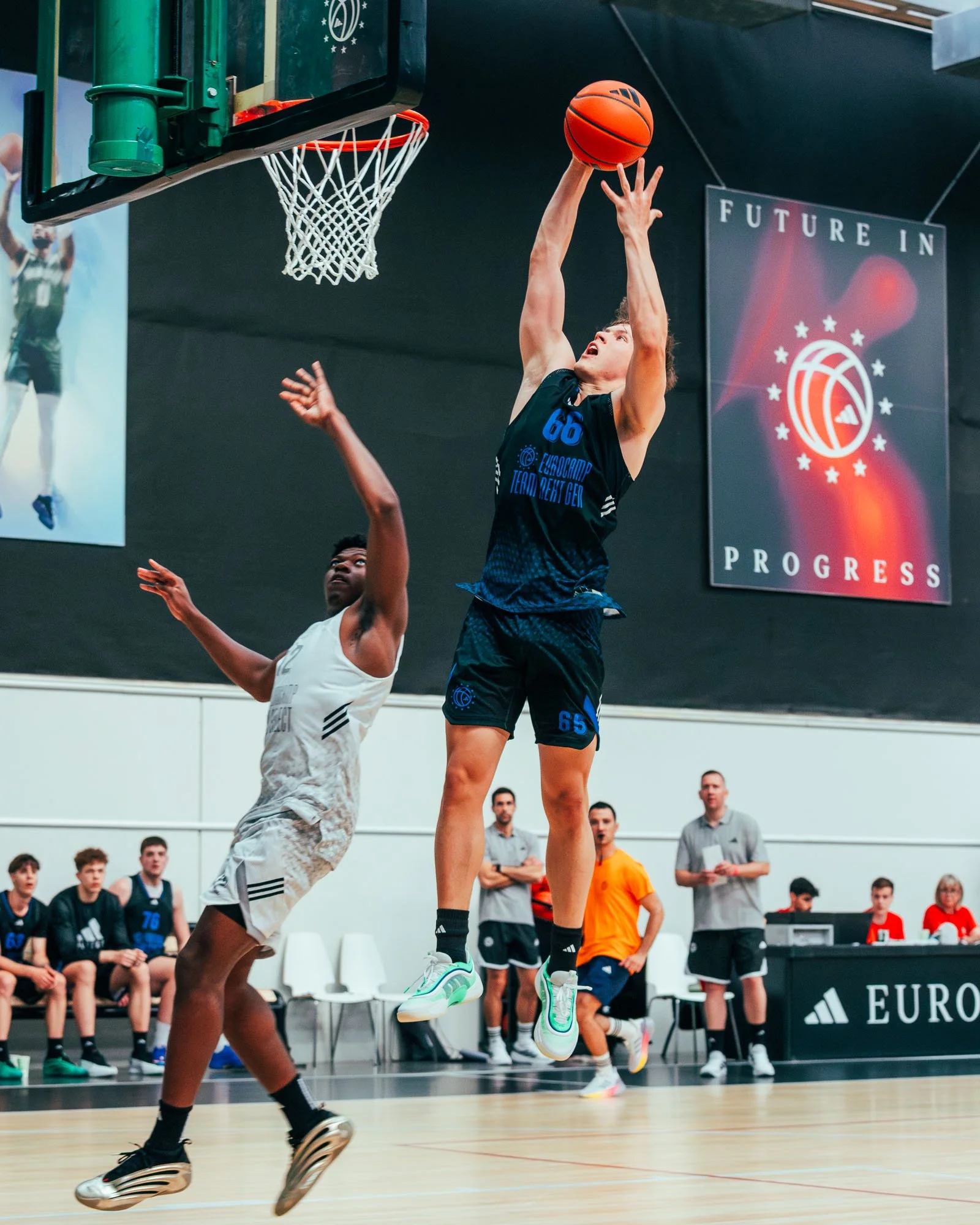 Giocatore di basket in aria mentre cerca di segnare un punto, con altri giocatori e pubblico sullo sfondo, in un campo da basket indoor