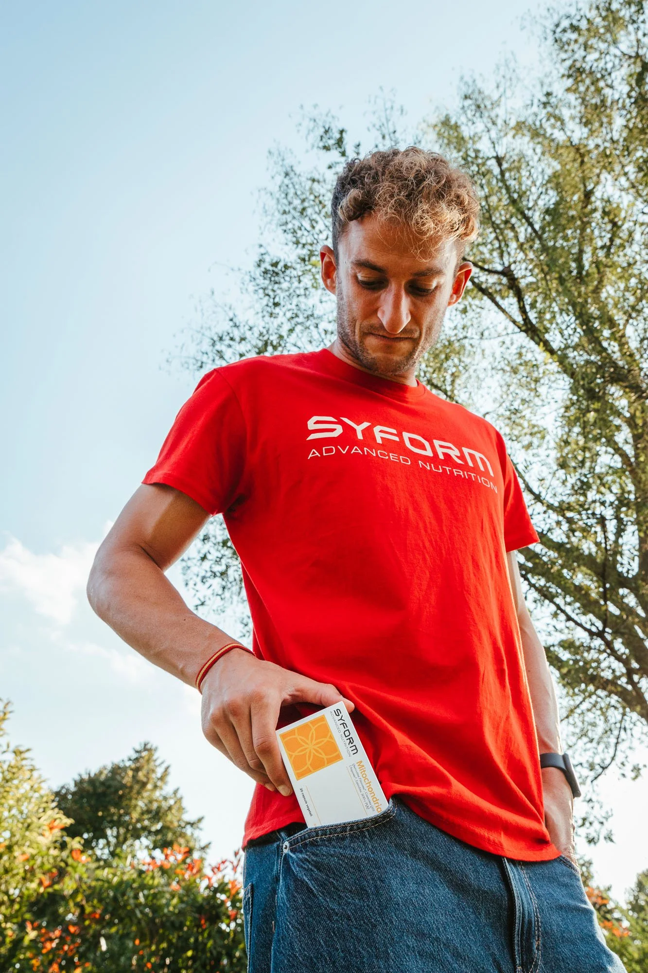 Un uomo con una maglietta rossa con scritto Syform Advanced Nutrition tiene una scatola di integratori, con uno sfondo di alberi e cielo chiaro.