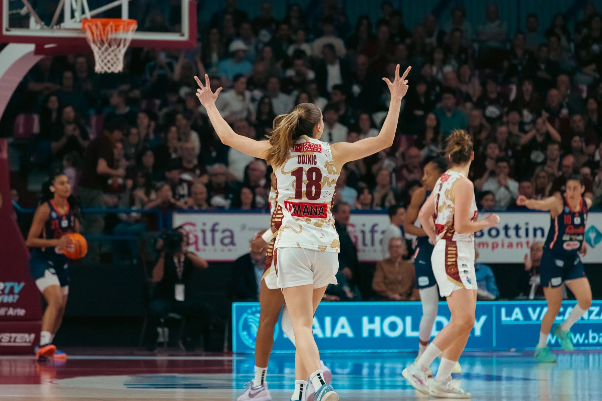 Giocatrice di basket femminile con capelli castani lunghi legati in coda, che indossa una maglia bianca con dettagli dorati e il numero 18, mentre fa un gesto decorativo con le mani in campo durante una partita. Sullo sfondo ci sono altre giocatrici,