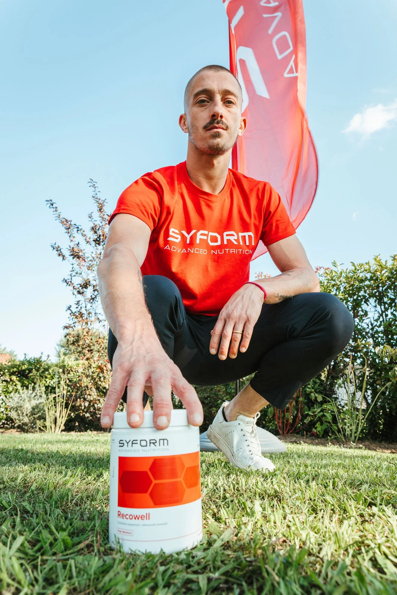 Un uomo con capelli rasati e maglietta rossa seduto sull'erba in giardino include in primo piano una scatola di integratore alimentare SYFORM Recowell. Sopra di lui, una bandiera rossa con logo SYFORM. Il cielo è azzurro e ci sono arbusti sullo sfond