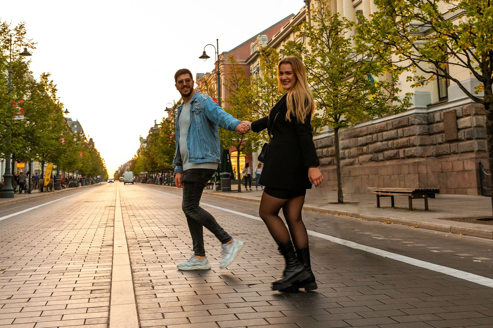 Due persone, un uomo e una donna, si tengono per mano mentre ballano o posano sulla strada di una città. La scena si svolge durante il tramonto, con alberi che laminano le facciate degli edifici e luci pubbliche accese.