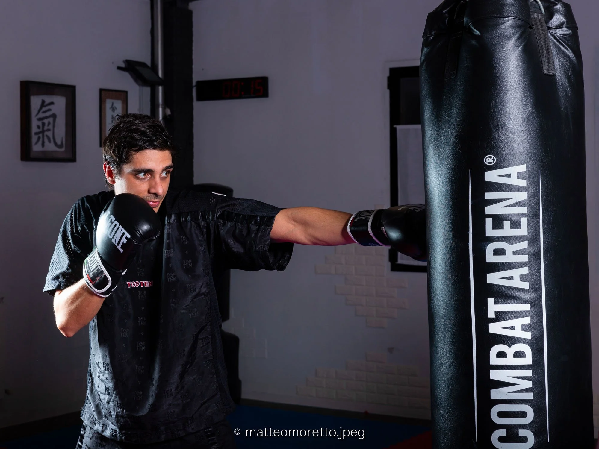 Un uomo che sta facendo esercizi di kickboxing in un centro di allenamento, colpendo un sacco da boxe. Indossa guanti da boxe e abbigliamento sportivo nero con dettagli grigi.