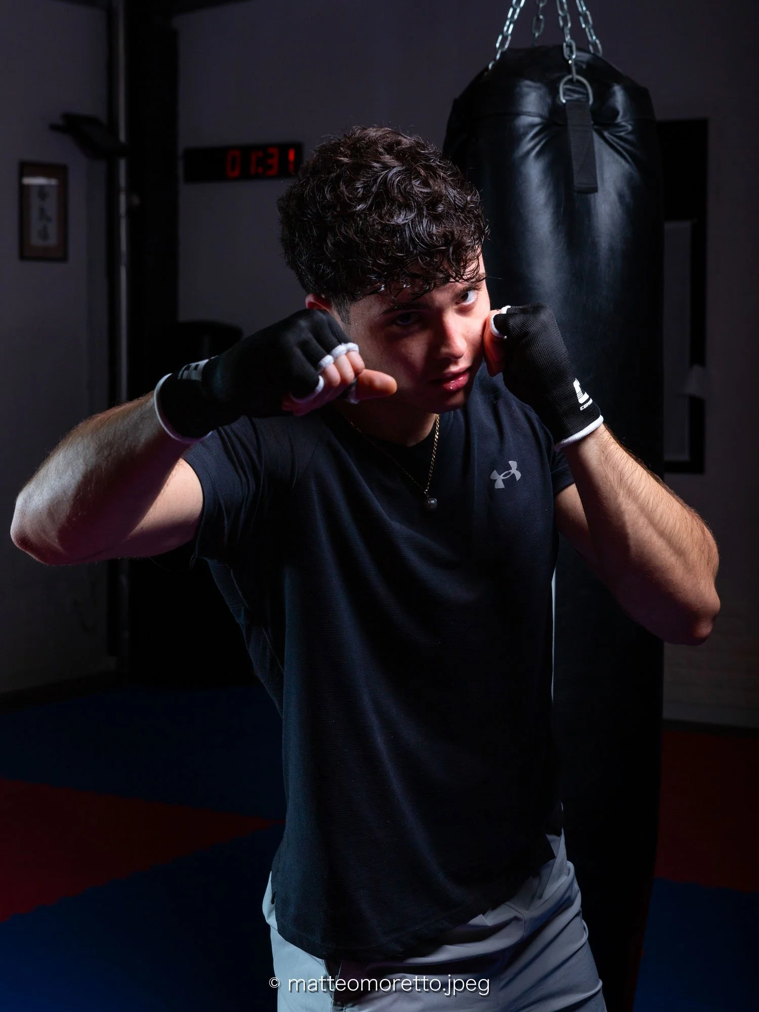 Giovane uomo con guantoni da boxe in posizione di combattimento in una palestra con sacco da boxe sospeso sullo sfondo. Luce focalizzata sul soggetto, sfondo scuro.