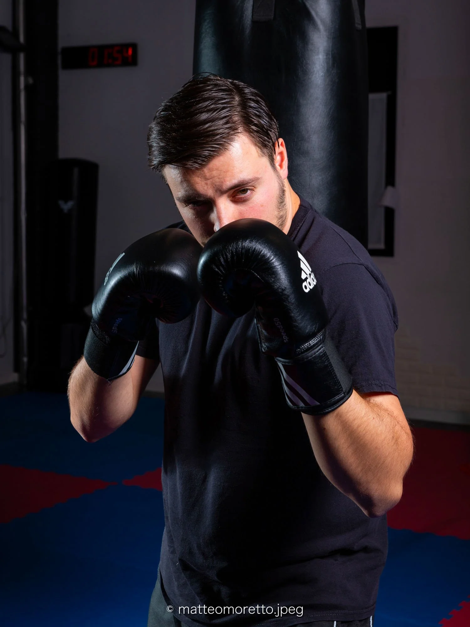 Uomo giovane con capelli castani scuri, in tinta unita nera, che si allena in palestra con guantoni da boxe e si mostra in posizione di combattimento di boxe davanti a un sacco da pugilato.