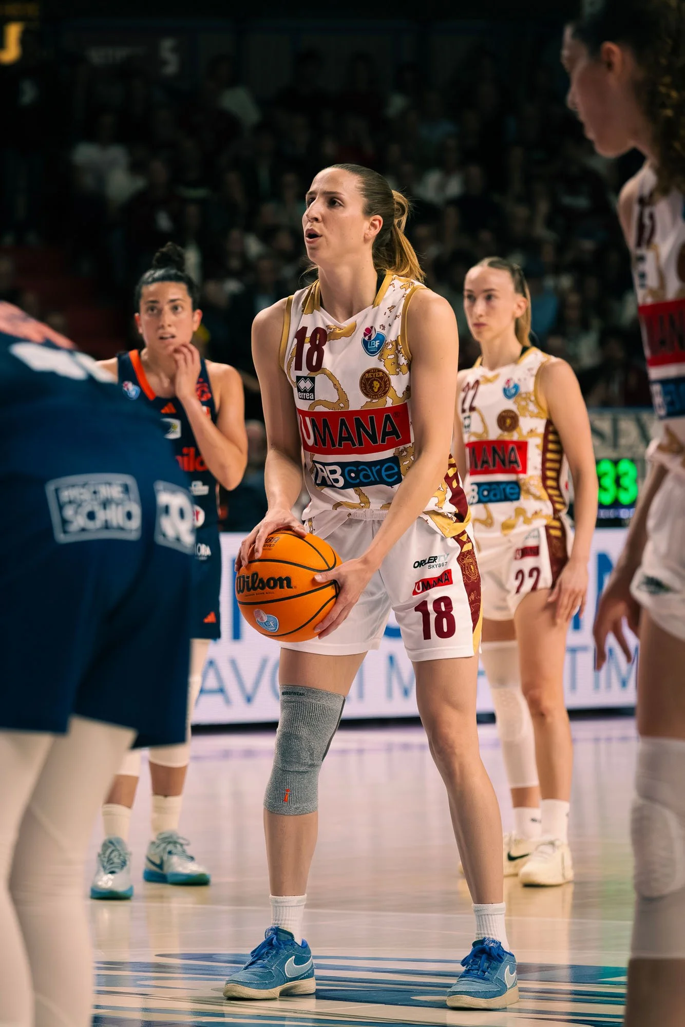 Giocatrice di pallacanestro con la palla in mano durante una partita, circondata da compagne in campo. Indossa un uniforme bianco con dettagli dorati e il numero 18.