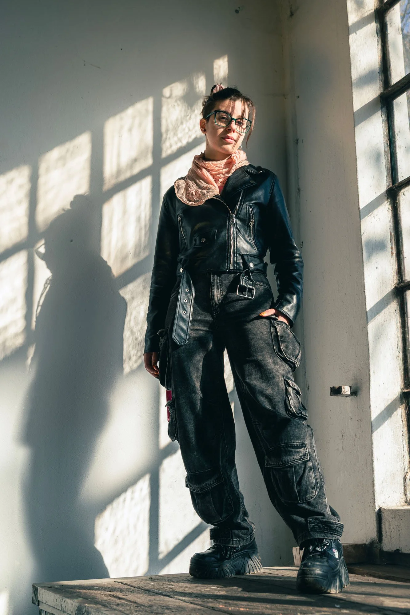 Ragazza con occhiali, giacca di pelle e pantaloni cargo neri, che si trova in uno spazio con parete bianca, illuminato dalla luce naturale attraverso una finestra con barre di metallo. La ragazza scatta un selfie o guarda verso la fotocamera, con un'