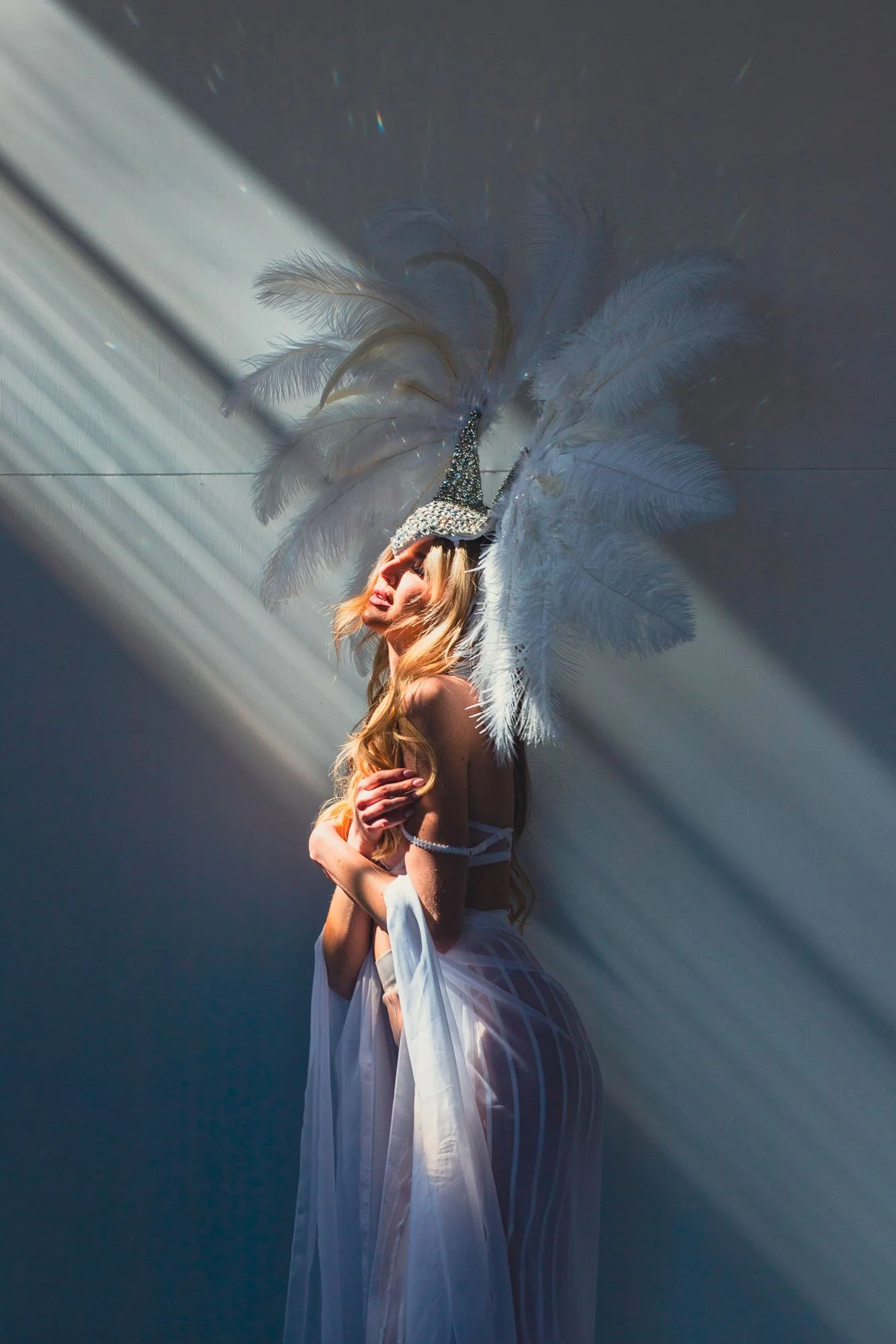 Donna con abito bianco, cappello decorato con piume e nastri, circondata da raggi di luce, posa con le braccia incrociate
