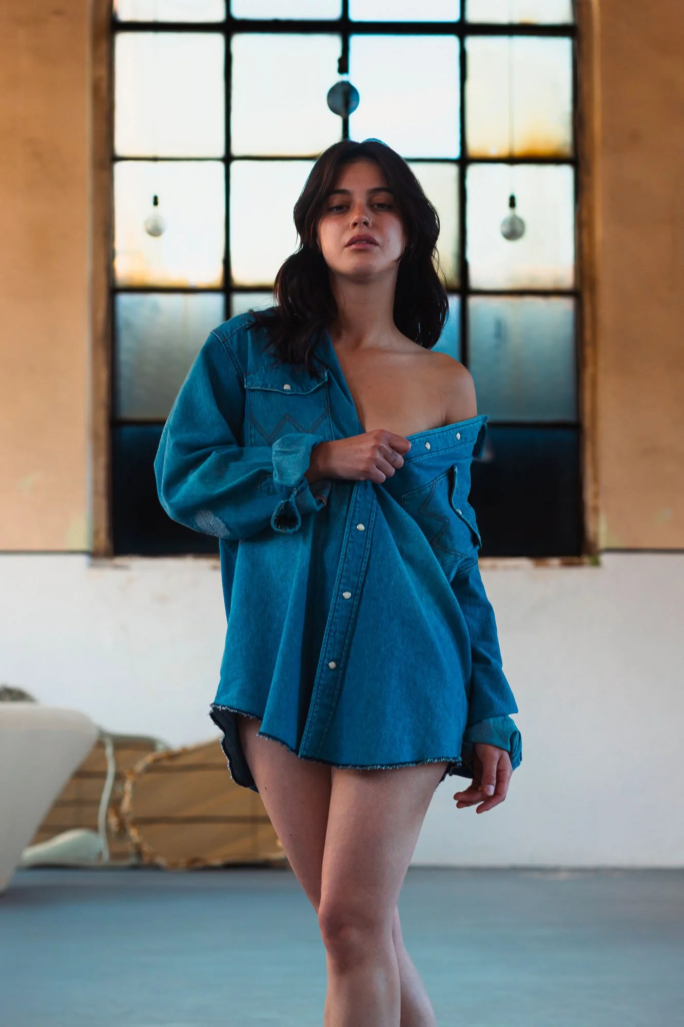 Una donna con capelli castani scuri, vestita con una giacca di jeans che indossa scarpe da ginnastica nere e shorts di jeans neri, si trova in un ambiente interno con pareti beige e una grande finestra sullo sfondo. La stanza ha alcuni arredi, tra cu