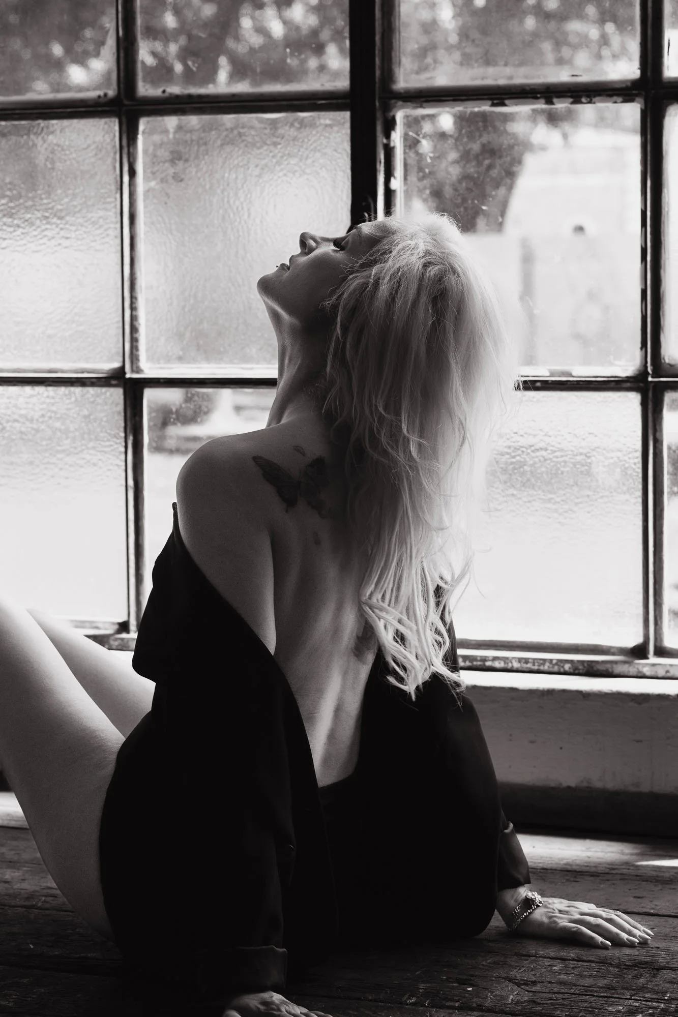 Una donna dai lunghi capelli biondi e tatuaggi, seduta per terra davanti a una grande finestra con vetri opachi, si inclina all'indietro con la testa e le braccia poggiate sul pavimento in legno.