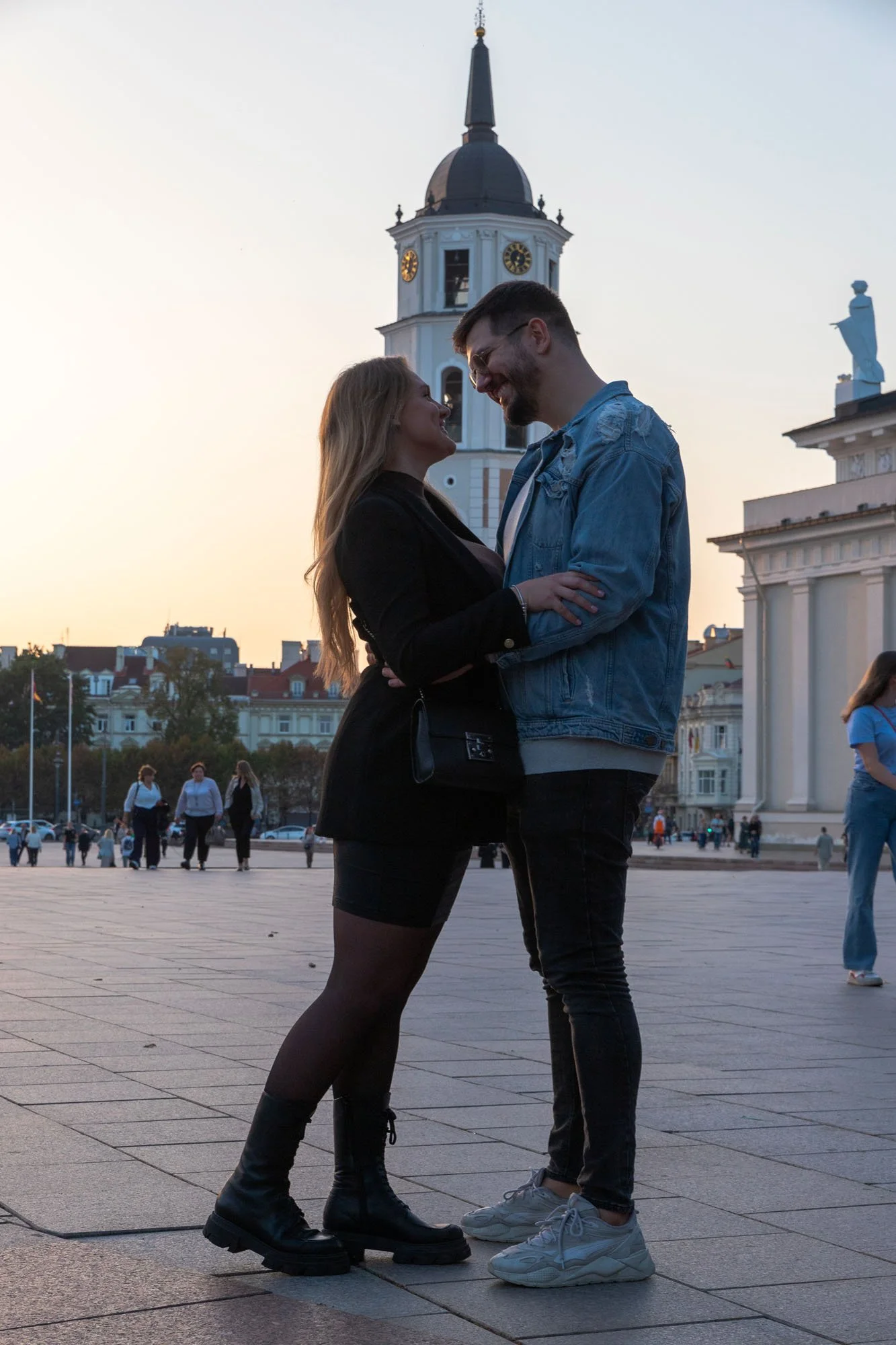 Una coppia si tiene per mano e si guarda negli occhi in una piazza al tramonto, con una chiesa e statue sullo sfondo.