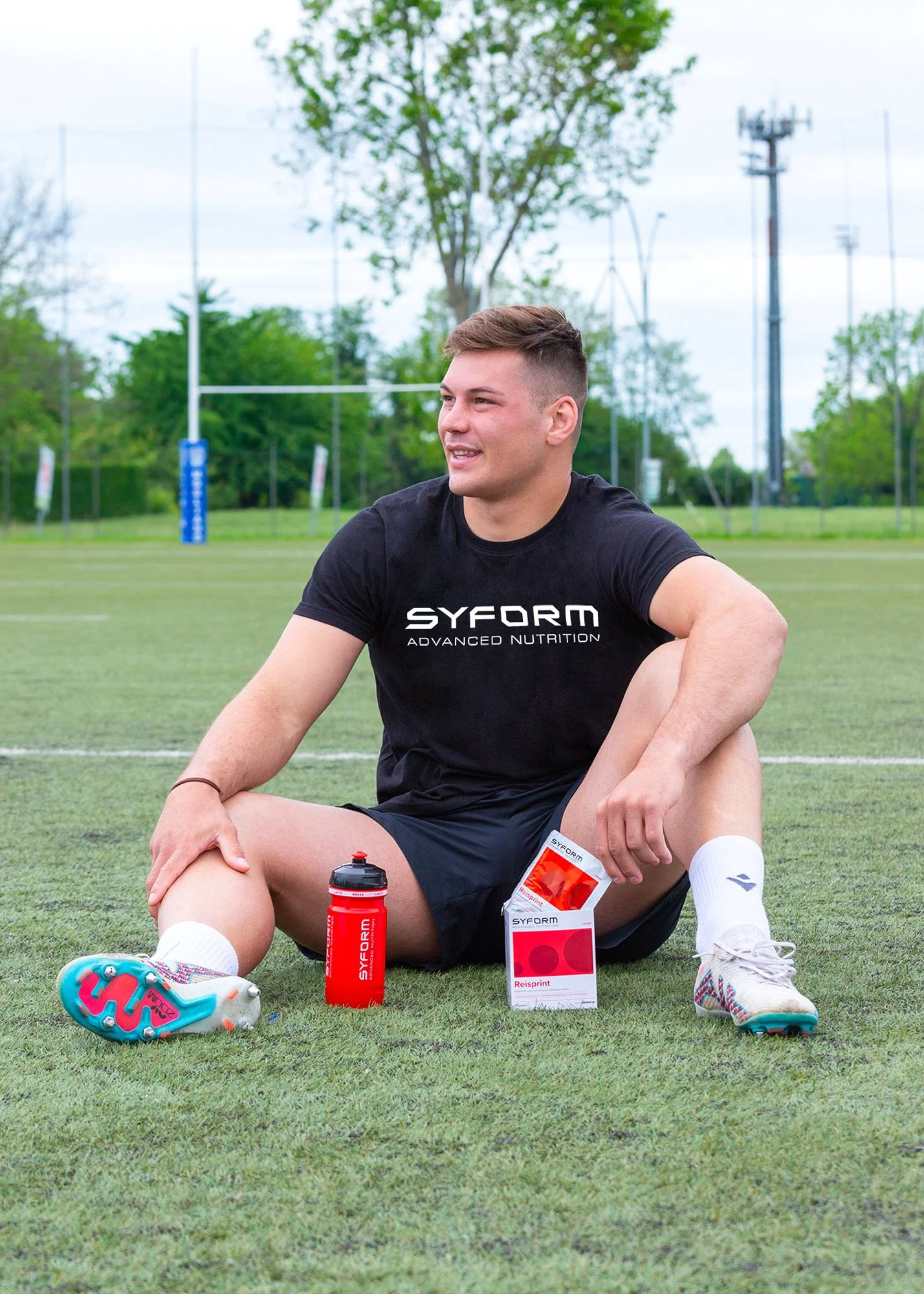 Giocatore di rugby seduto sul campo con supplementi alimentari e acqua vicino a lui, indossa maglietta nera con scritto 'SYFORM ADVANCED NUTRITION'.