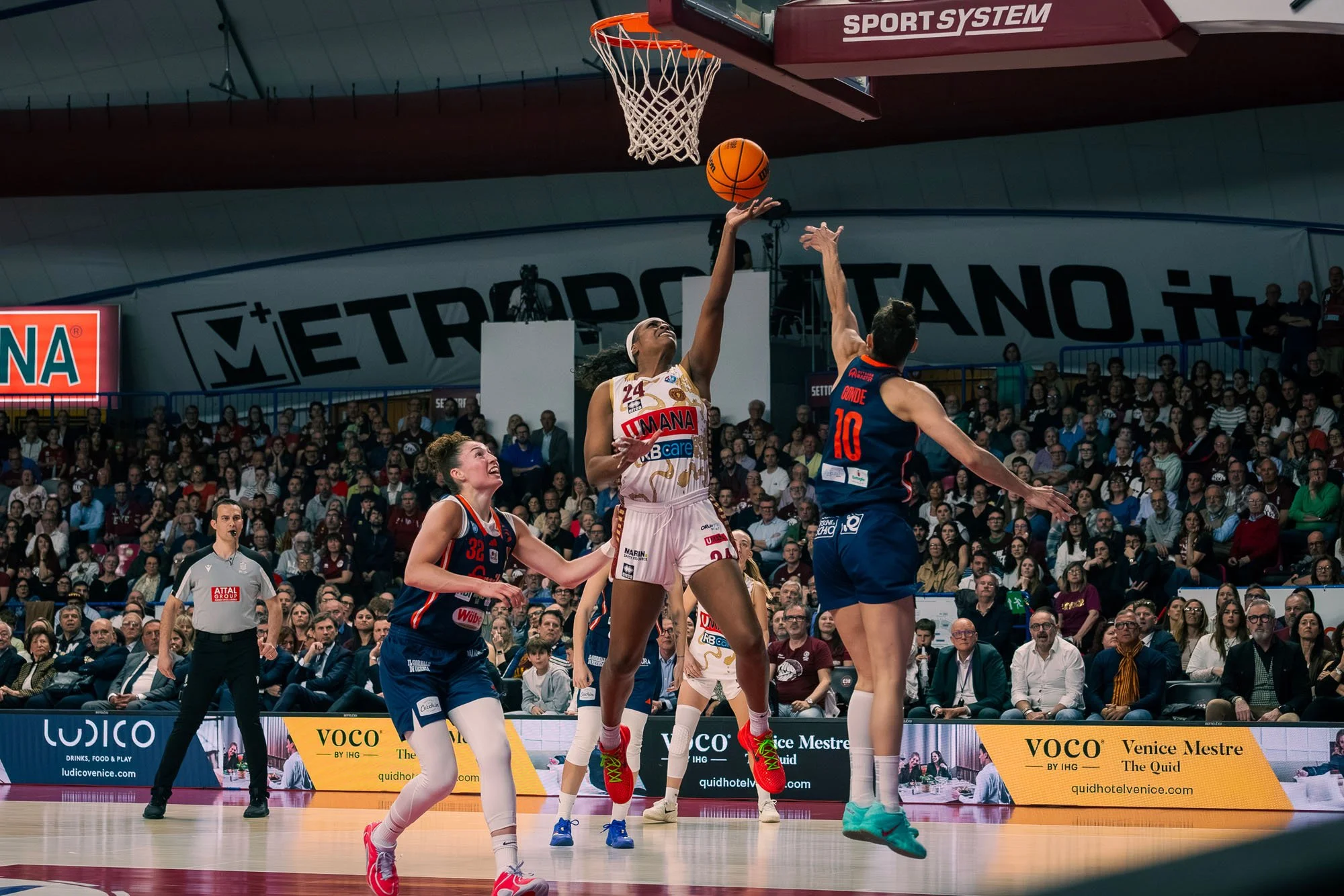 Giocatrici di pallacanestro in azione durante una partita in un'arena affollata, con pubblico seduto e un arbitro che osserva.