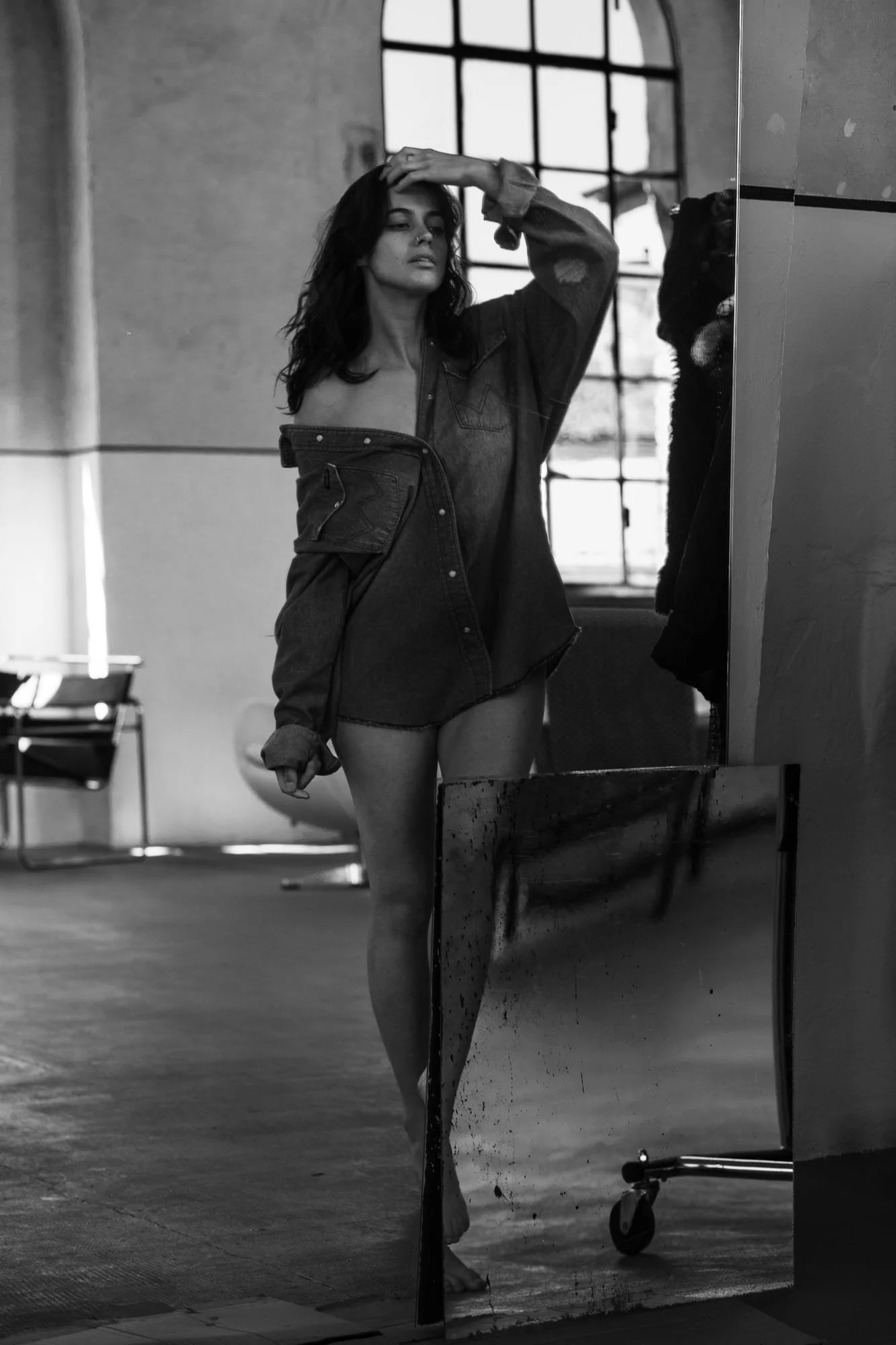 Ragazza in posa davanti a uno specchio in uno studio fotografico, vestita con una giacca aperta. Immagine in bianco e nero.