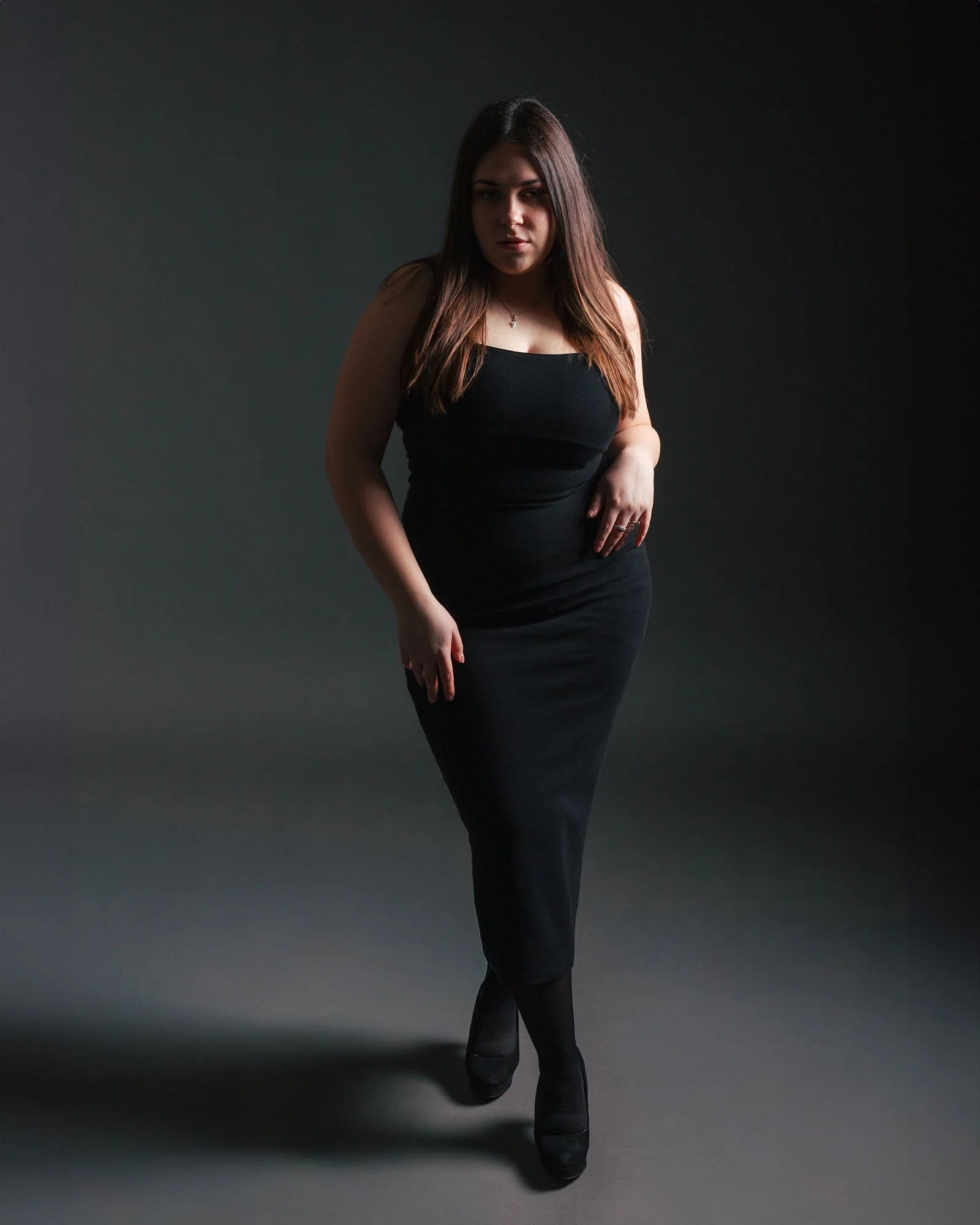 Ragazza vestita di nero, nera in piedi su sfondo scuro, con braccia rilassate, luci drammatiche su e giù, look elegante e sguardo intenso.
