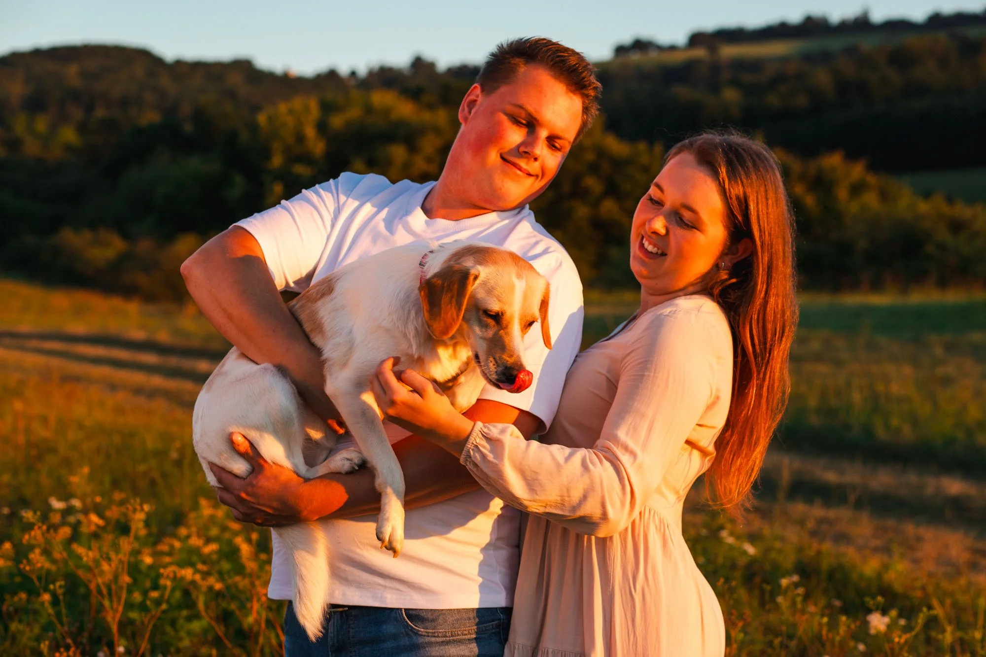 Una coppia sorridente abbraccia un cane bianco e marrone durante il tramonto in un campo verde.