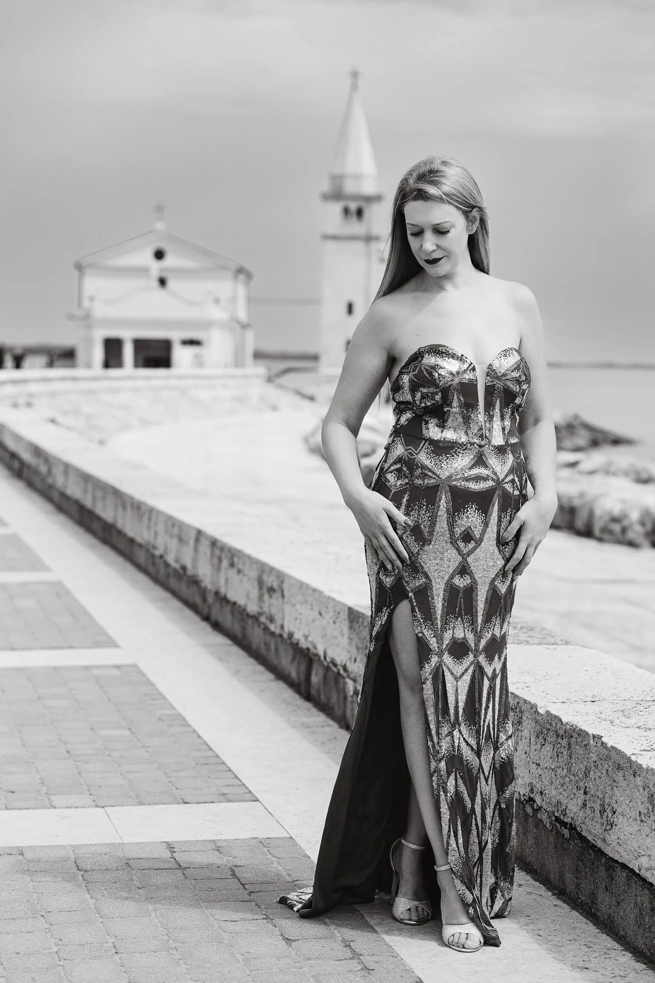 Una donna in abito lungo con scollo a cuore a stampa geometrica si trova sul lungomare, con edifici e una torre sullo sfondo, in bianco e nero.