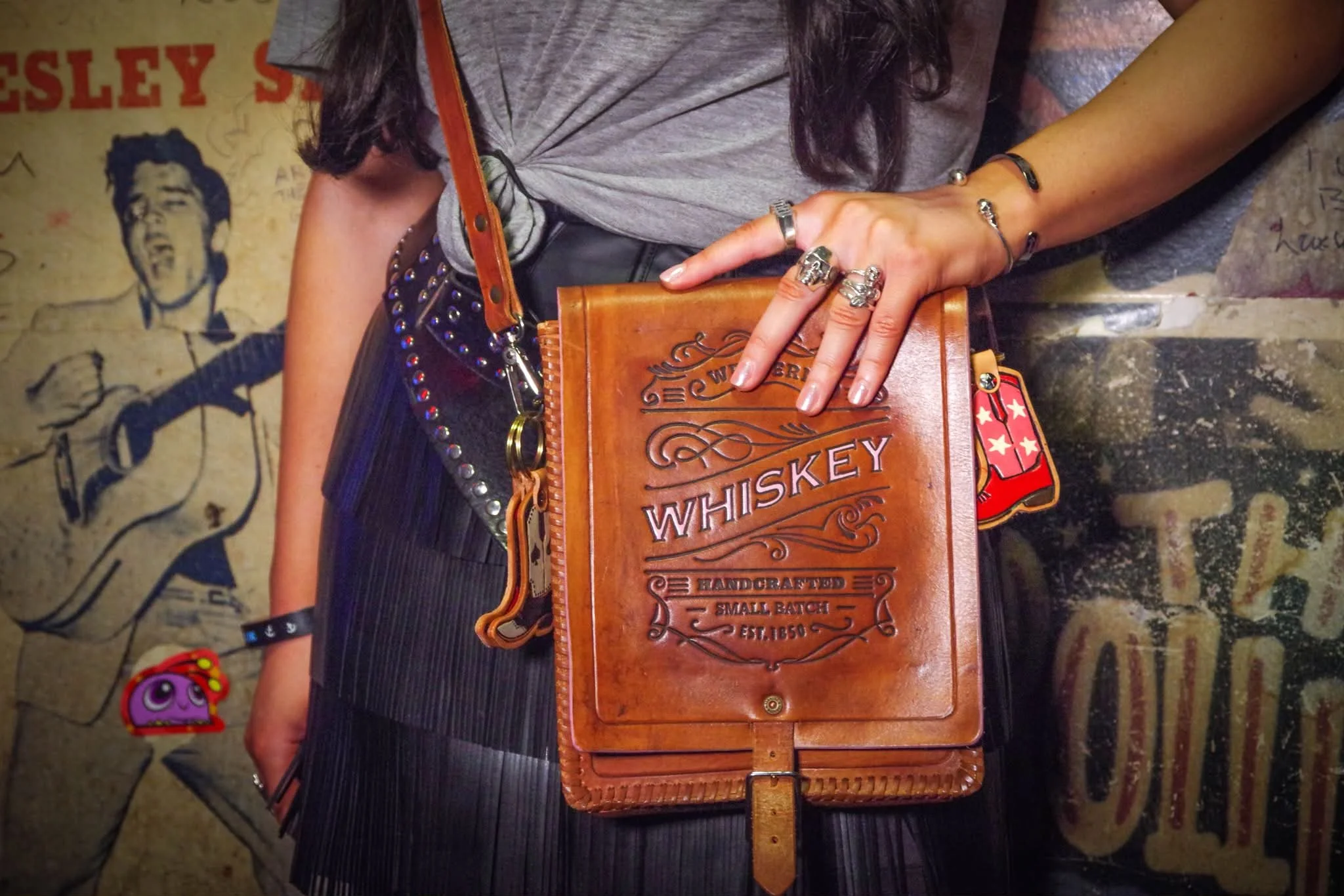 Person wearing handcrafted tan leather Whiskey satchel bag by Dead Cowboy London in rock venue setting with cowboy boot keyring and silver rings.