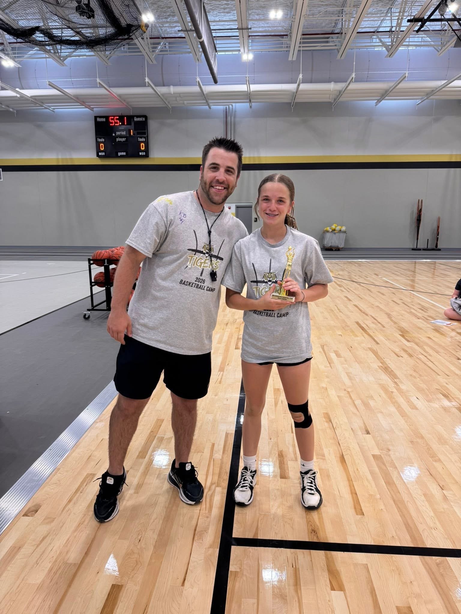 A man and young girl in a gymnasium, smiling and posing for a photo. The girl holds a small trophy, and both are wearing gray T-shirts with a sports logo. The man wears black shorts and sneakers; the girl wears black shorts, a knee brace, and sneakers.