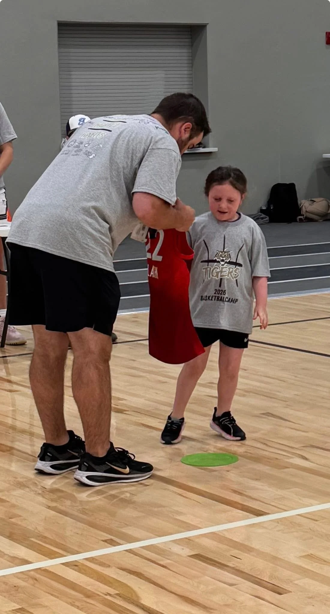 A young girl receiving instructions from an adult man on a basketball court indoors. The girl is wearing a gray t-shirt with 'TIGERS 2026 BASKETBALL CAMP' printed on it, and black sneakers. The man is dressed in athletic clothing, including a gray t-shirt, black shorts, and black sneakers, bending down to talk to her.
