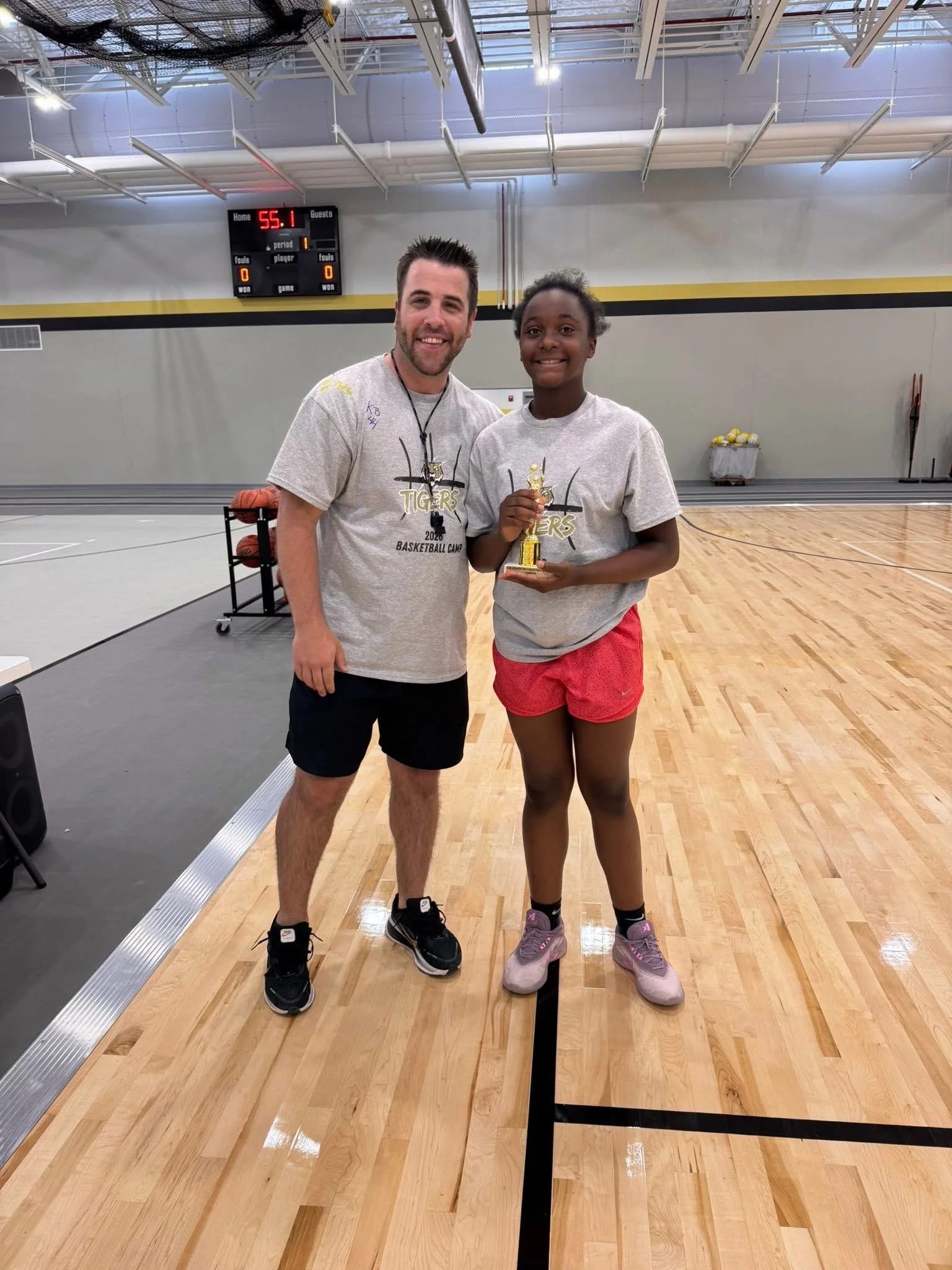 A young female basketball player holding a trophy, smiling, standing next to a male coach or trainer in a gymnasium with a scoreboard in the background.
