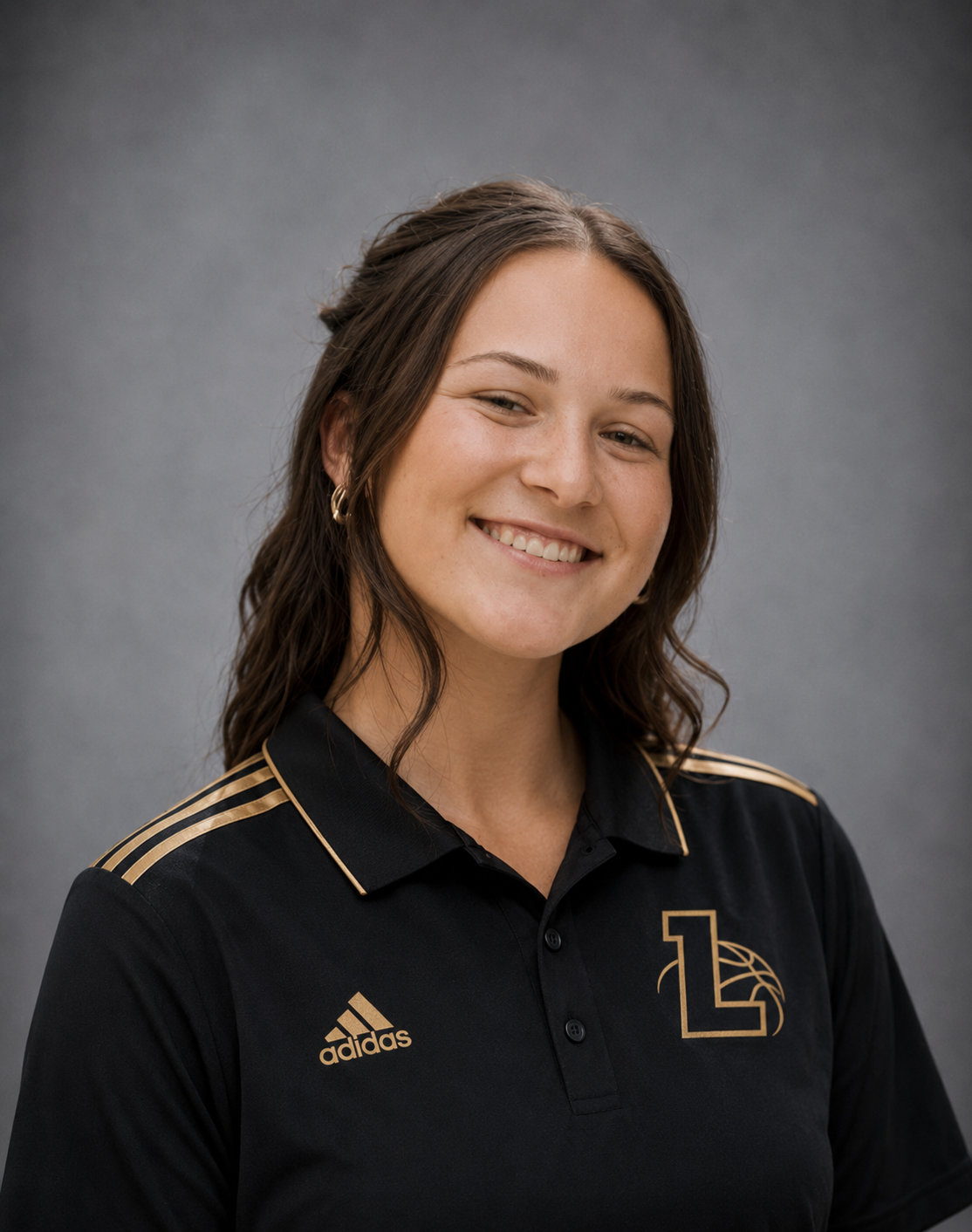 Smiling young woman with brown hair wearing a black Adidas sports polo with gold accents and a logo on the chest, standing against a gray background.