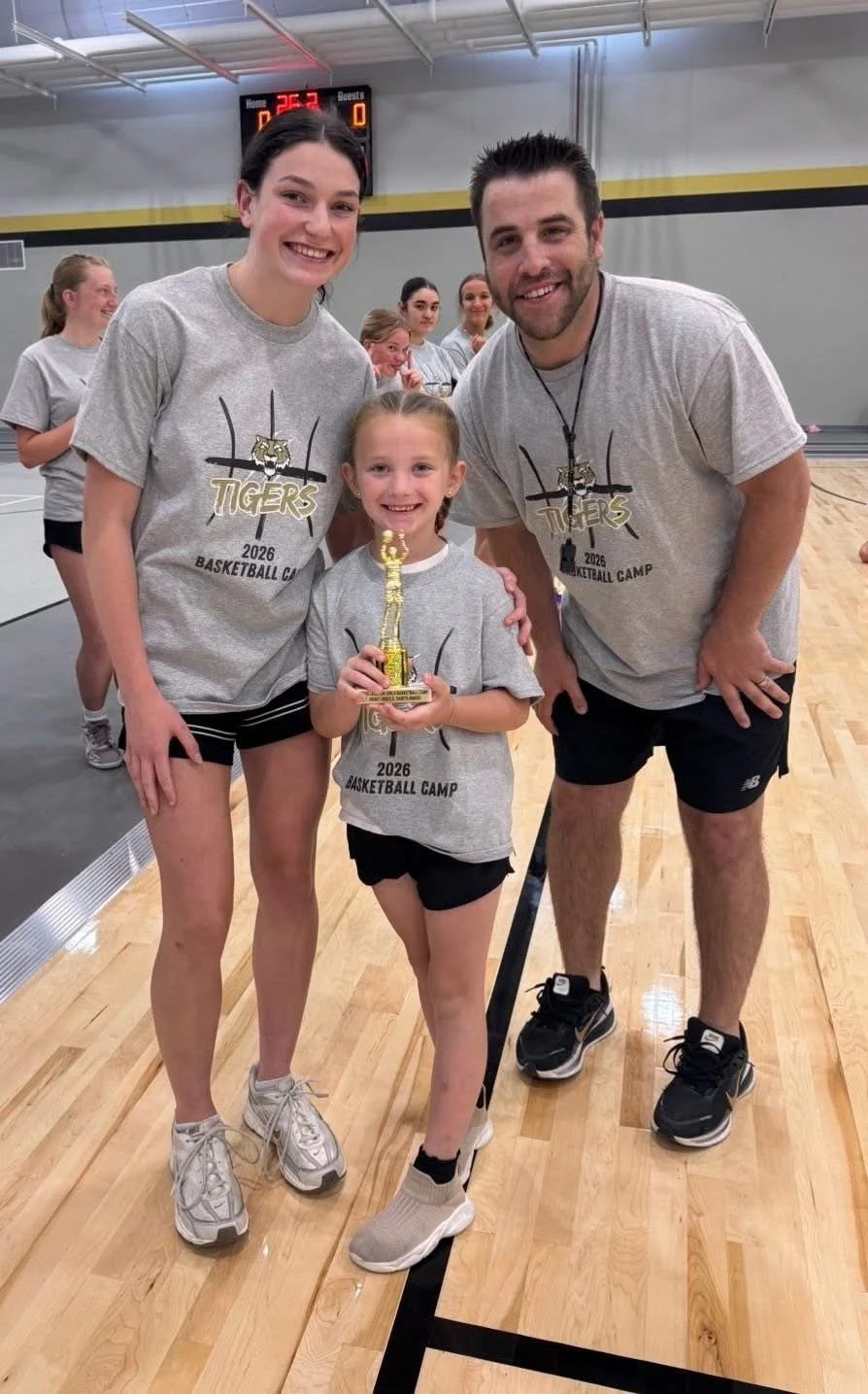 A young girl holding a trophy poses with two adults and other girls in a gymnasium. They are all wearing matching 'Tigers 2026 Basketball Camp' T-shirts, smiling after a sports event.
