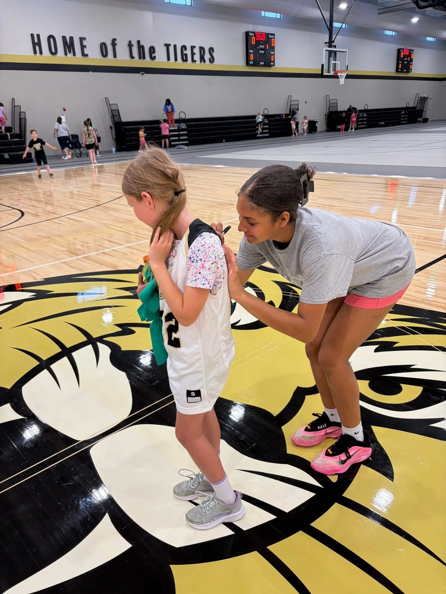 A girl with a ponytail and sporting shoes stands on a basketball court with a woman with braids, bending down and talking to her. The girl wears a sports jersey, shorts, and sneakers. The woman wears athletic clothing and bright pink sneakers. In the background, there are other children and a small audience on the bleachers at a gymnasium, with a sign that reads 'Home of the Tigers' and a digital scoreboard above the court.