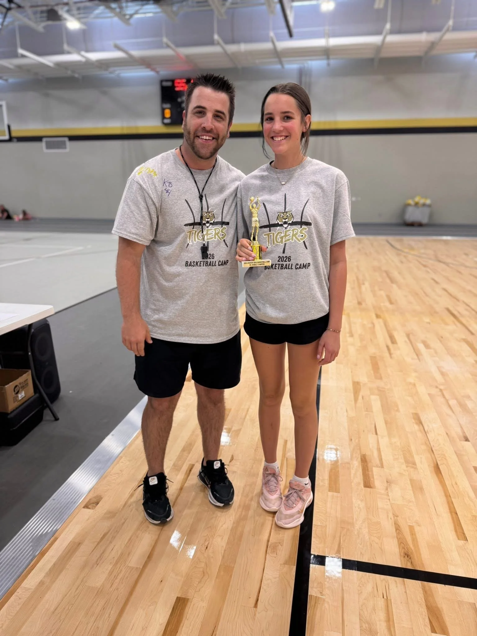 Two smiling young people, a man and a woman, standing on a basketball court, holding a trophy. Both are wearing gray T-shirts with 'Tigers Basketball Camp 2026' printed on them.