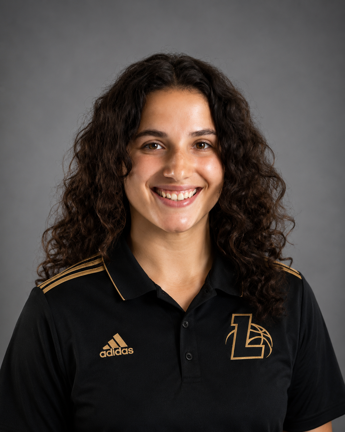A woman with curly brown hair wearing a black Adidas sports shirt with gold accents and a logo on the chest, smiling at the camera against a gray background.