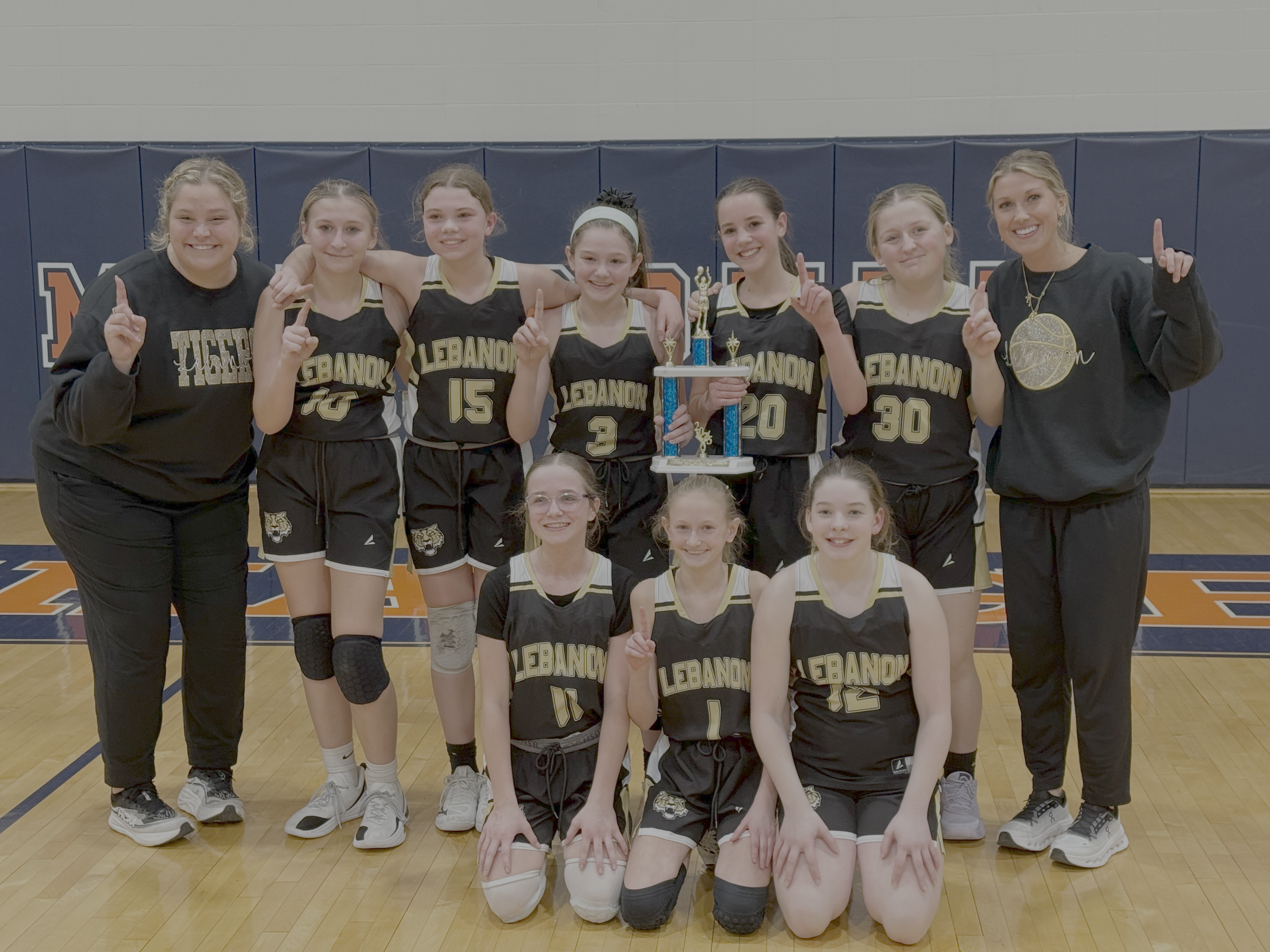 A girls basketball team is celebrating after a game, with some players and their coach smiling, and a trophy held by one of the players. They are in their black and gold uniforms in a gymnasium.