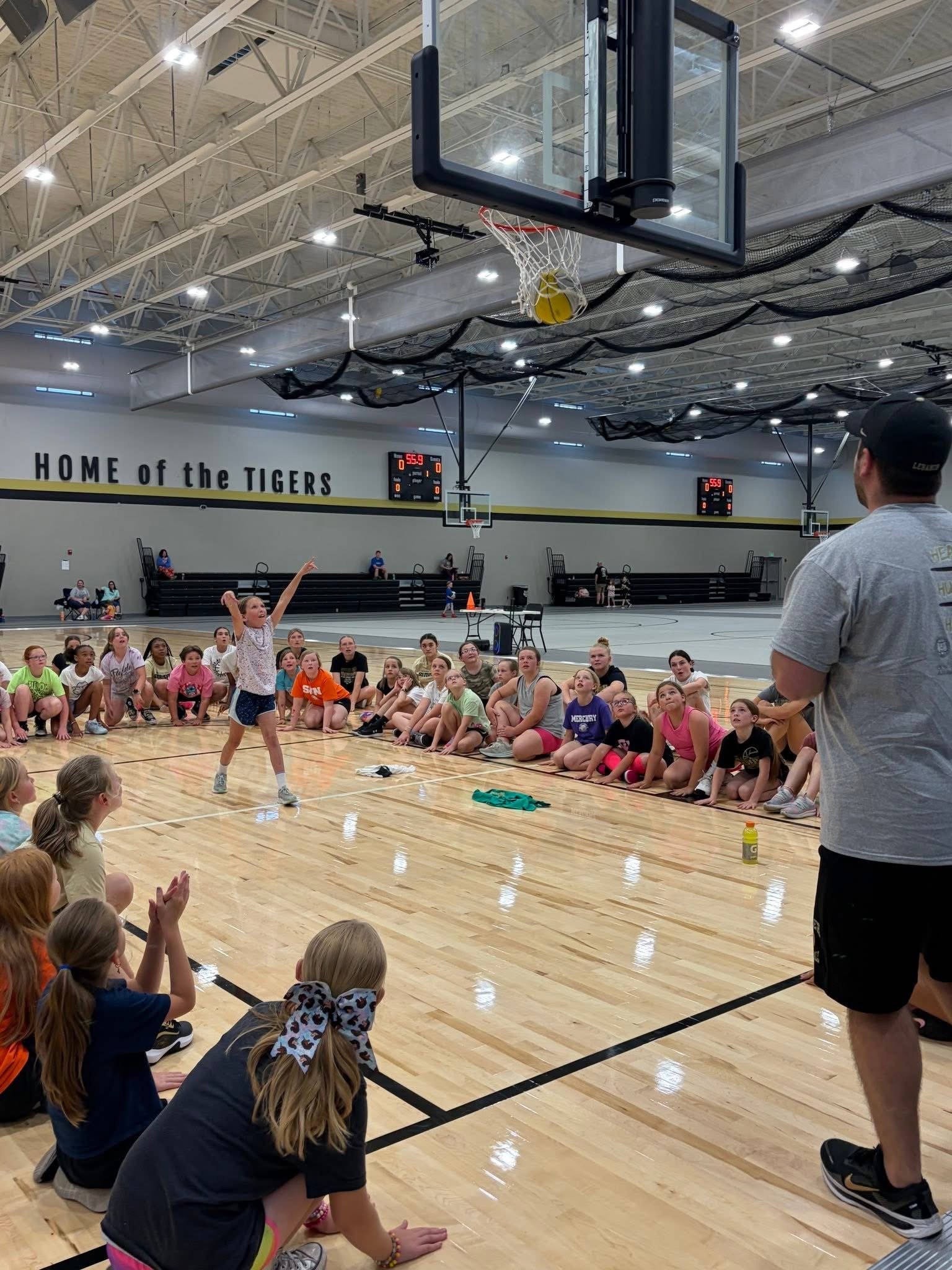 Children sitting on a gymnasium floor watching a girl attempt a basketball shot with a coach observing nearby.