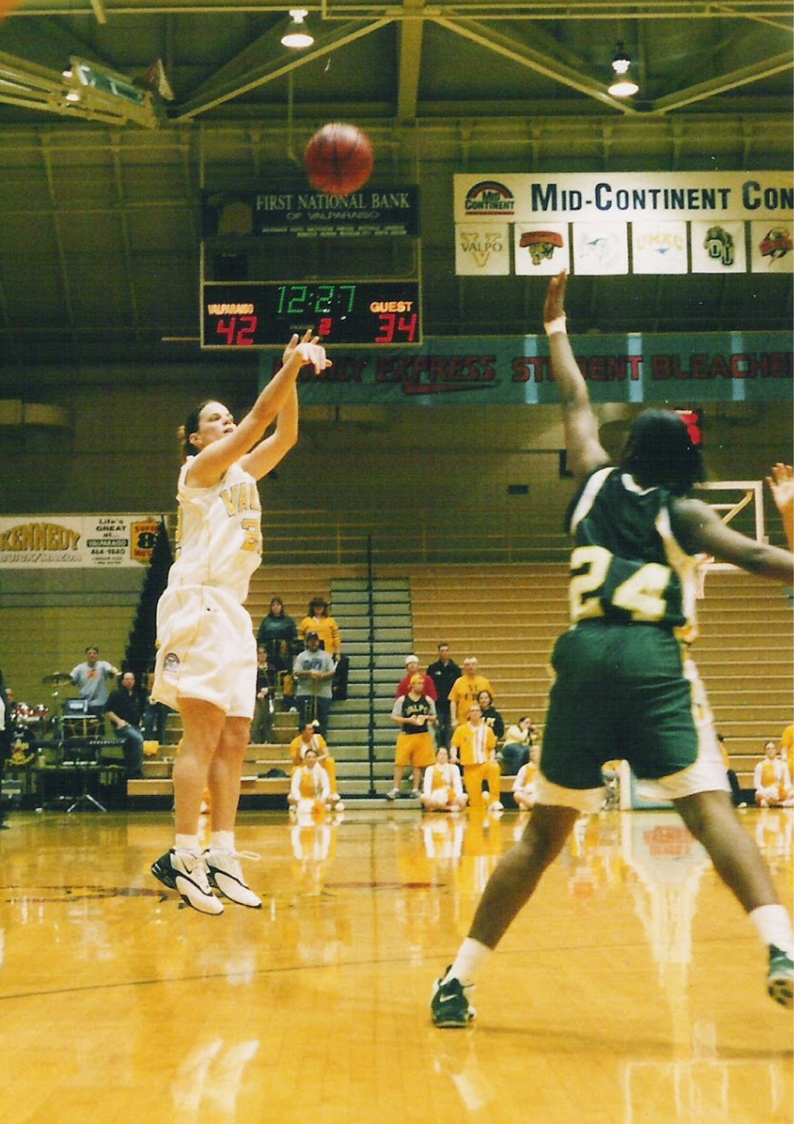 Women playing basketball on an indoor court with a scoreboard showing 42-34, and spectators and players in the background.