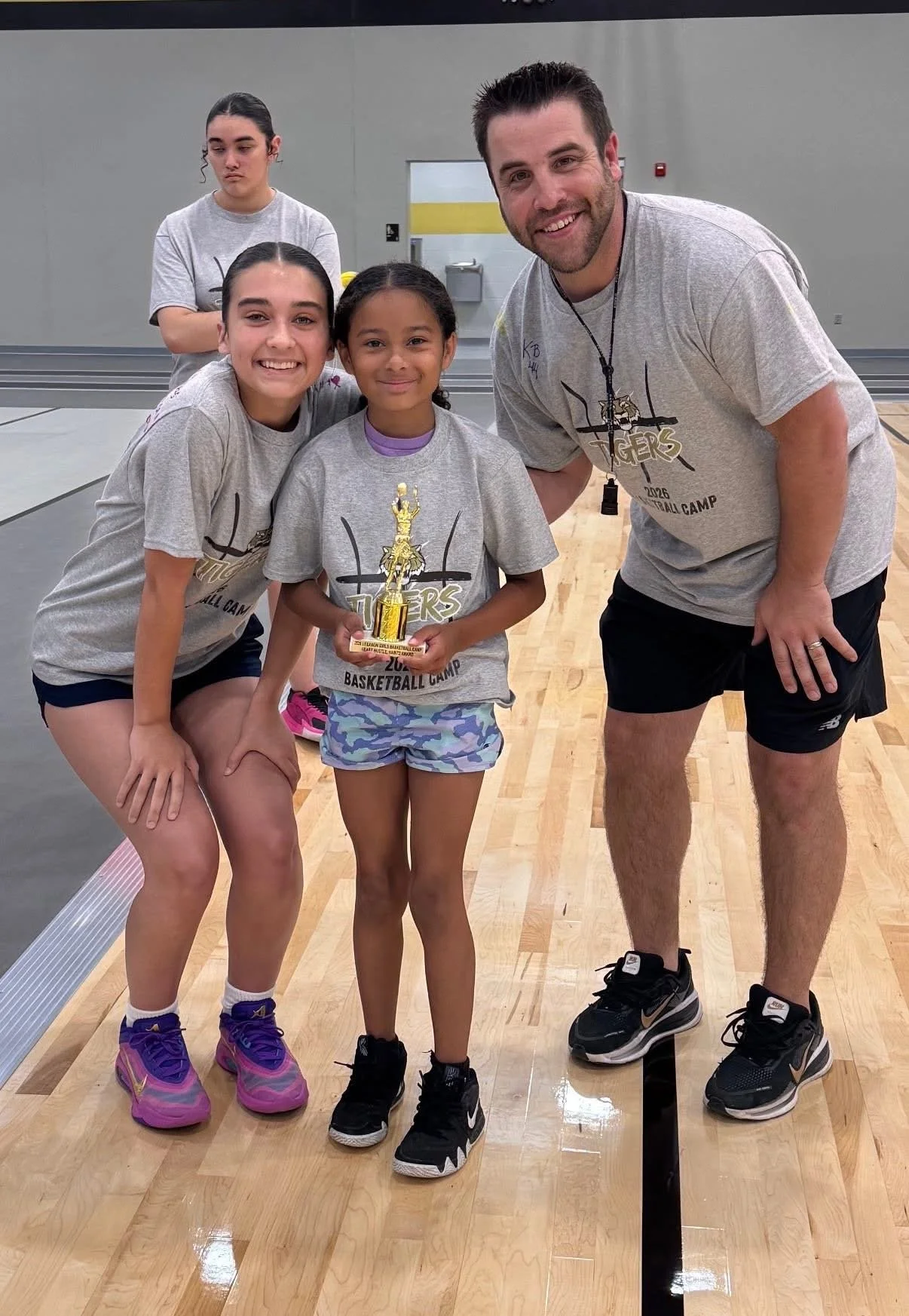 Girls and coach at basketball camp, one girl holding a trophy, in a gym with hardwood floors.