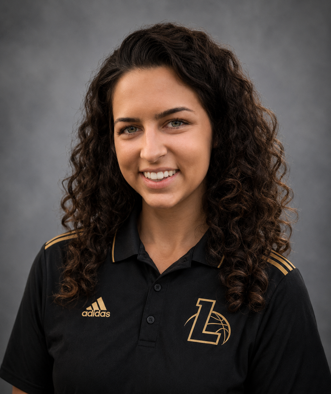 A young woman with curly dark hair, smiling, wearing a black Adidas polo shirt with gold accents and a sports logo on the chest.