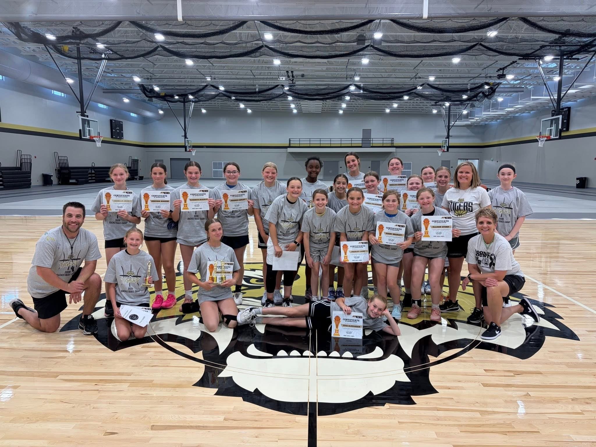 A group of young female basketball players and their coaches inside a basketball court, holding certificates and trophies, smiling for the photo.