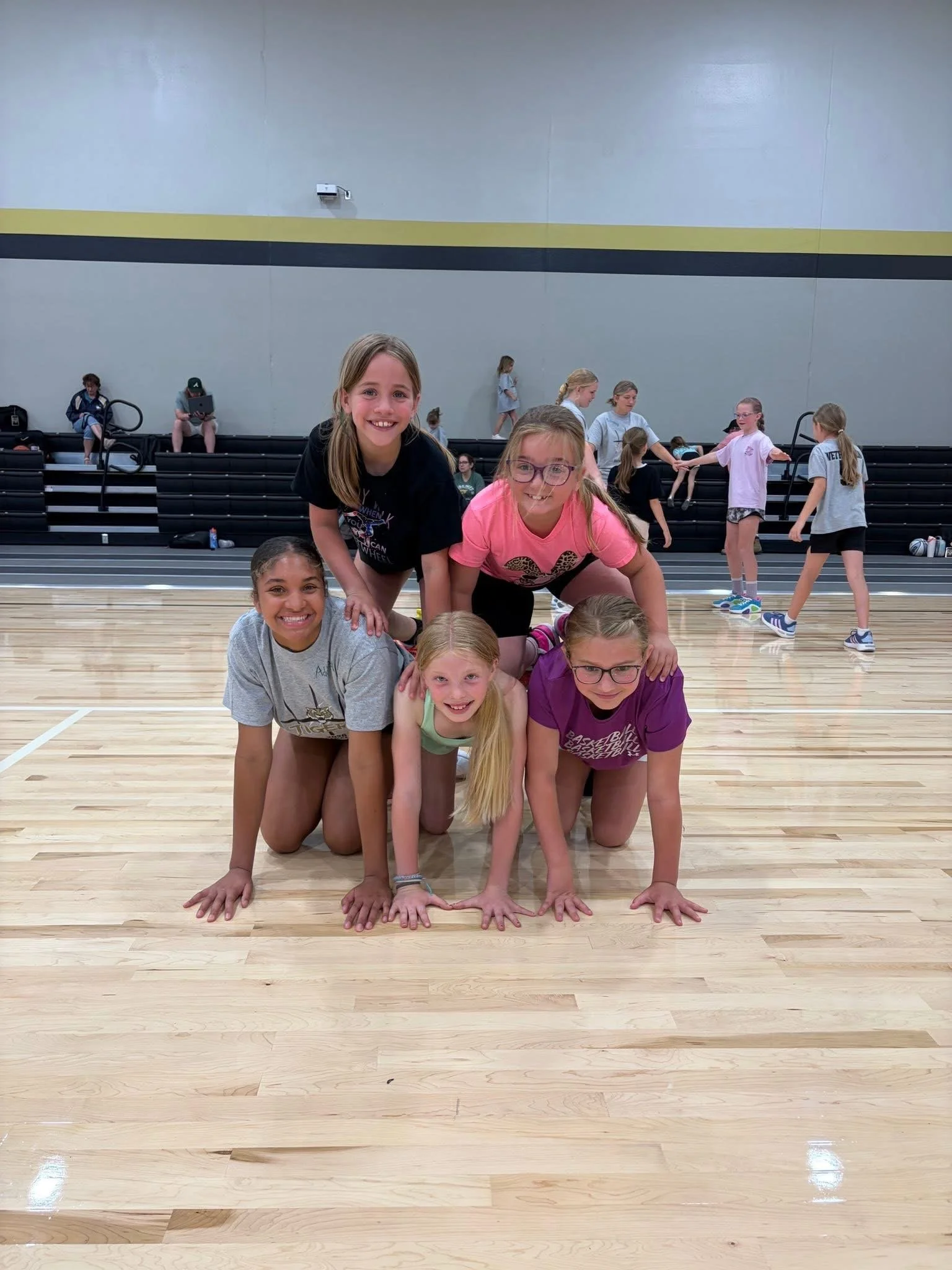 A group of young girls posing on gymnasium floor, with some girls supporting others in a pyramid, others walking or sitting in the background.