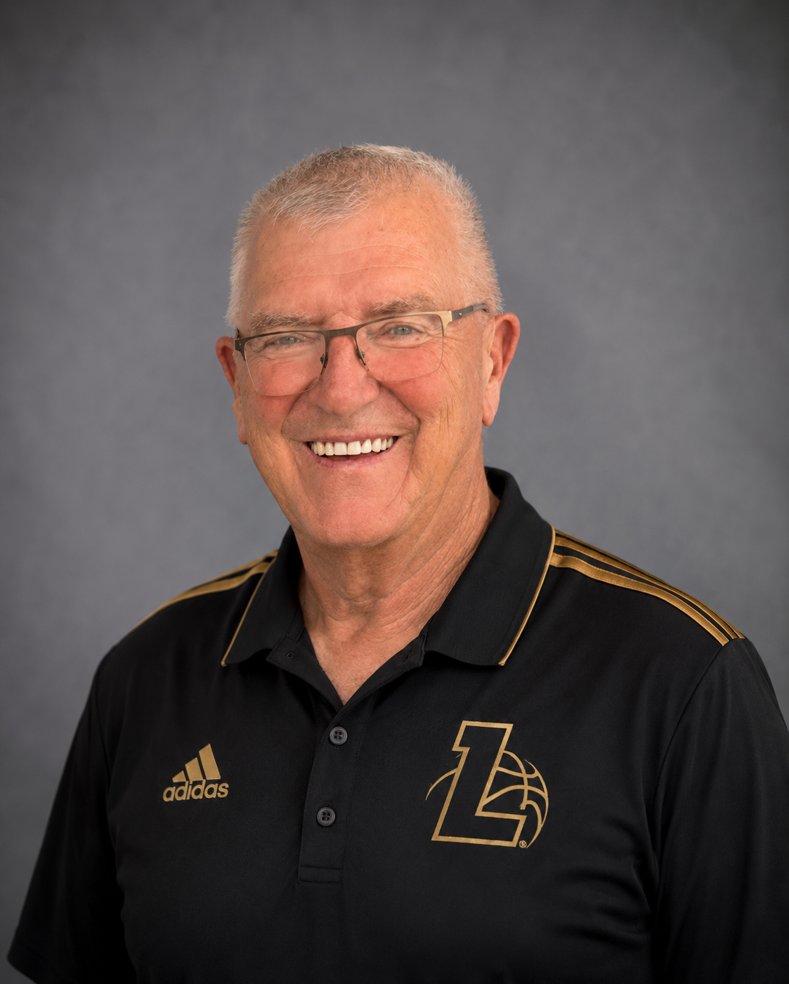 A smiling man with gray hair and glasses wearing a black Adidas shirt with gold accents and a logo with a 'L' and a basketball on the chest, posing against a gray background.
