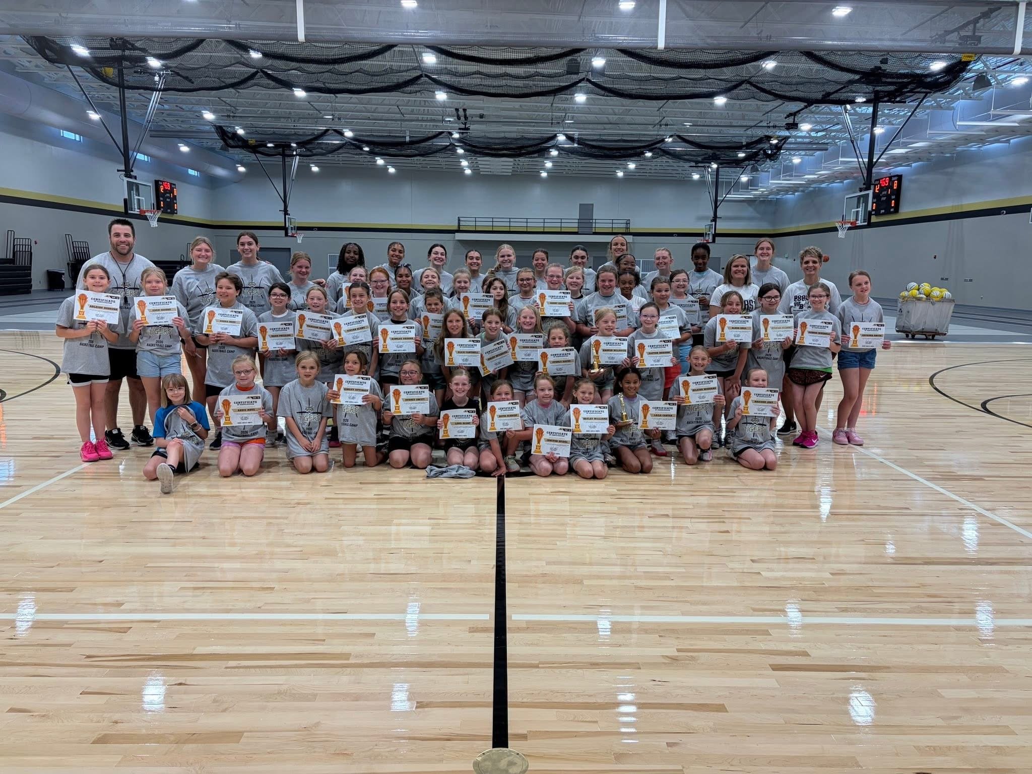 A large group of young girls and coaches in a gymnasium, holding certificates after a basketball camp, smiling for the photo.