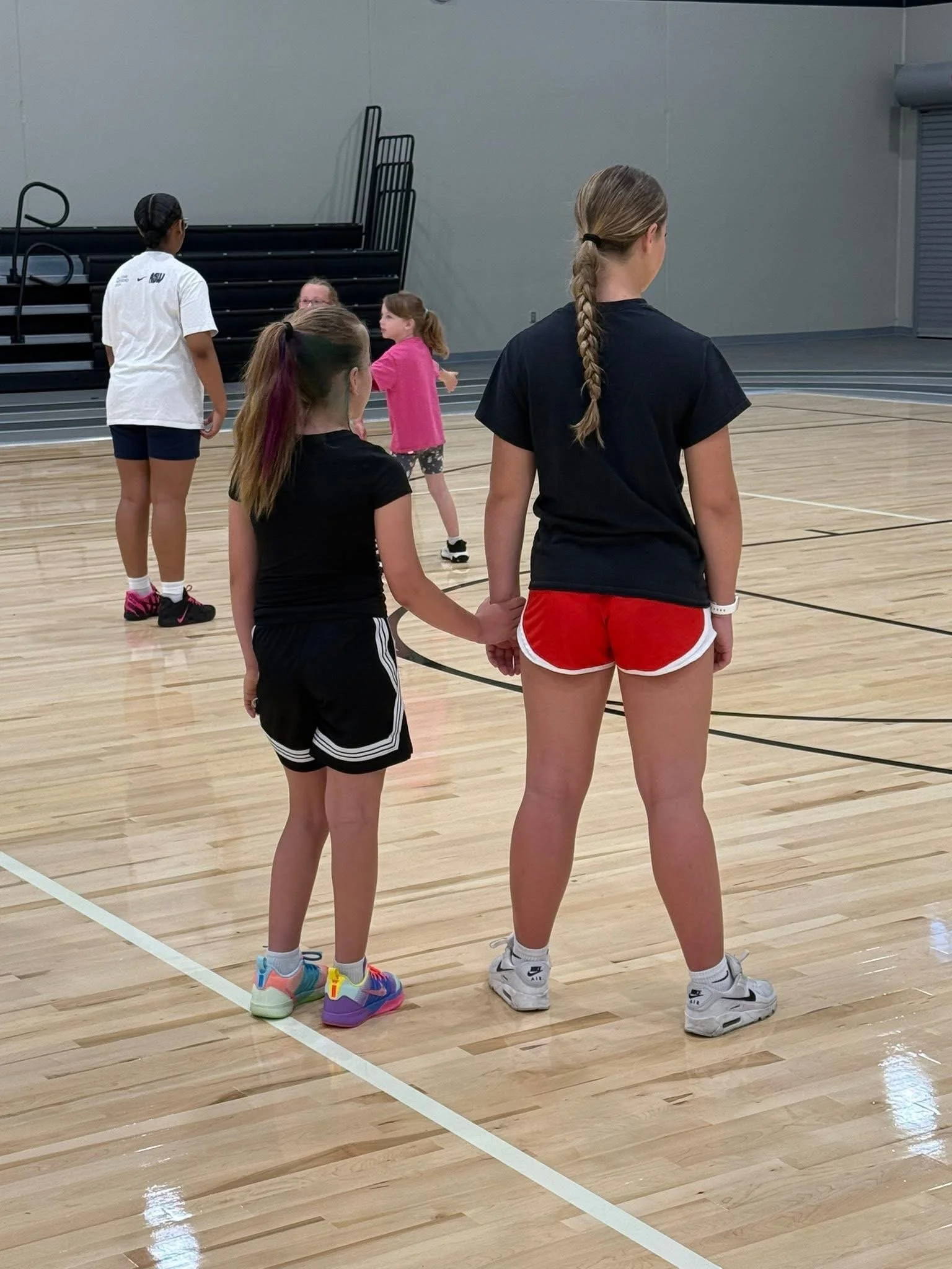 Four girls in sports attire standing on a basketball court, facing away, with one girl holding hands with another. They are in a gym.