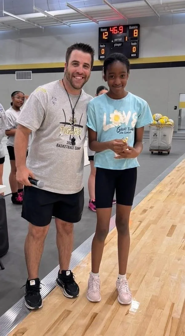 A man and a young girl standing inside a gymnasium, smiling at the camera. The man is wearing a gray t-shirt and black shorts, and the girl is wearing a light blue t-shirt and black shorts. Behind them, a scoreboard shows a score of 12 to 8 with 46.9 seconds remaining. The girl is holding an object in her hands.