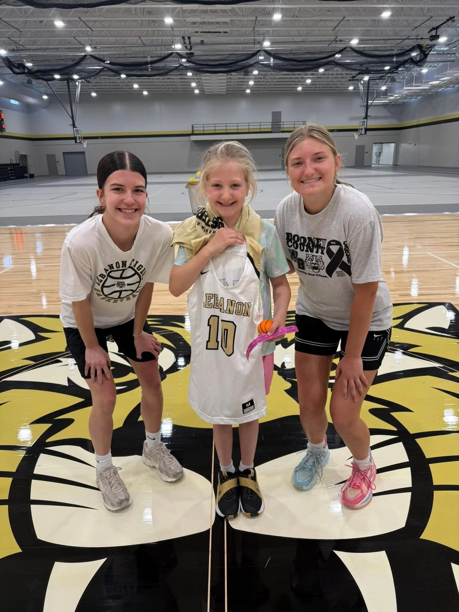 Three young girls standing on a gymnasium basketball court, smiling at the camera. The girl in the middle is holding a basketball with a gold ribbon around her neck. The background shows gymnasium walls, basketball hoops, and a mirror.