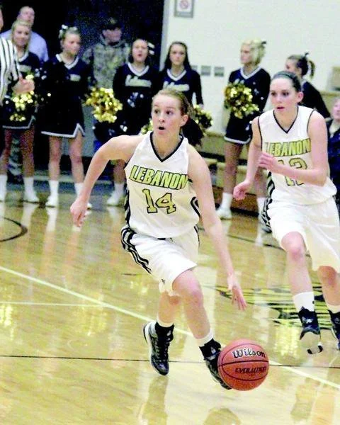 Two female basketball players in white Lebanon uniforms playing on a wooden court, with cheerleaders in black uniforms and pom-poms in the background.