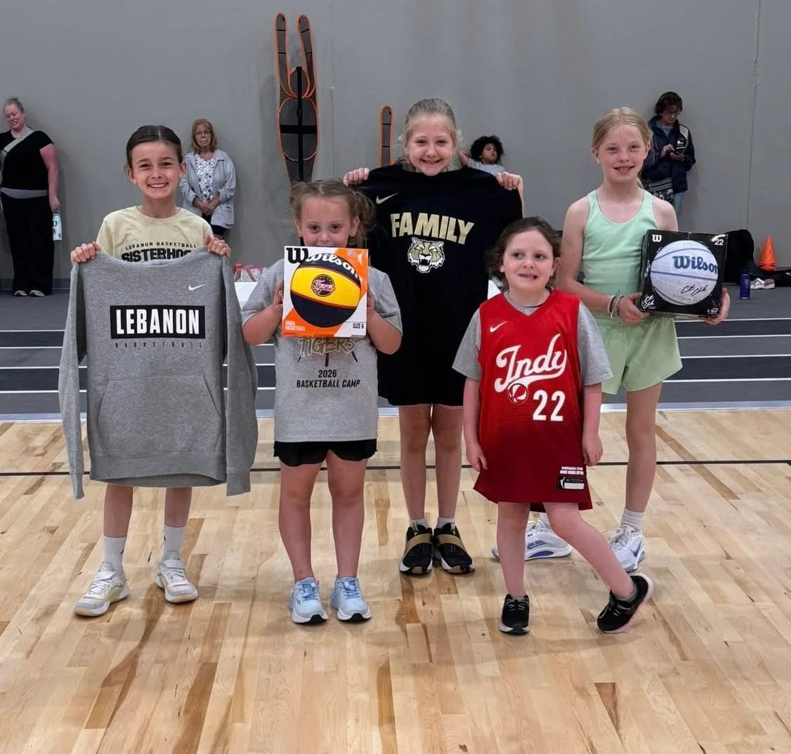 Group of young girls in a gymnasium holding sports apparel and volleyballs, smiling at the camera, with a wall featuring an eagle and a basketball in the background.