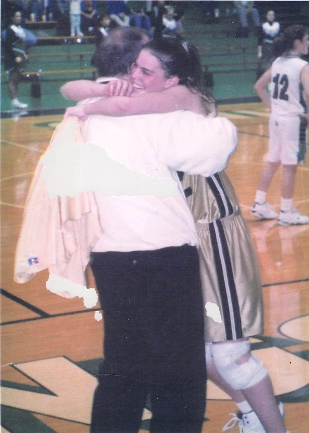 Two people hugging on a basketball court, with basketball players in the background.