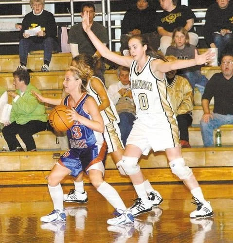 Two female basketball players, one in a blue uniform with the number 3 and the other in a white uniform with the number 0, competing on a basketball court surrounded by spectators.